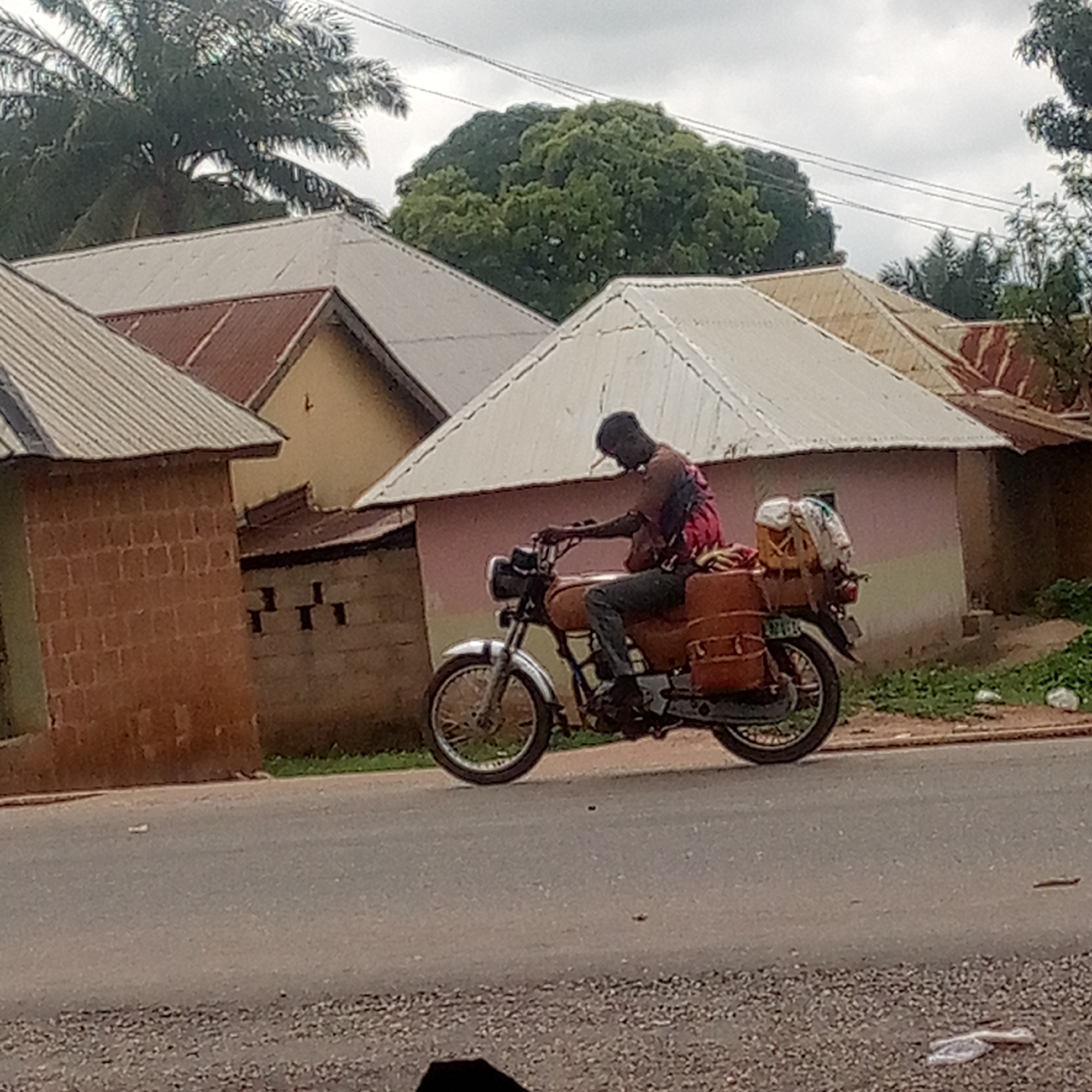 A man is riding a motorcycle on a paved road in Mararaba, Nigeria. The motorcycle has two brown leather saddlebags and is carrying a bundle of items secured on the back. The man is wearing dark pants and a red and black top. The background consists of buildings with corrugated metal roofs and trees. The sky is overcast.
