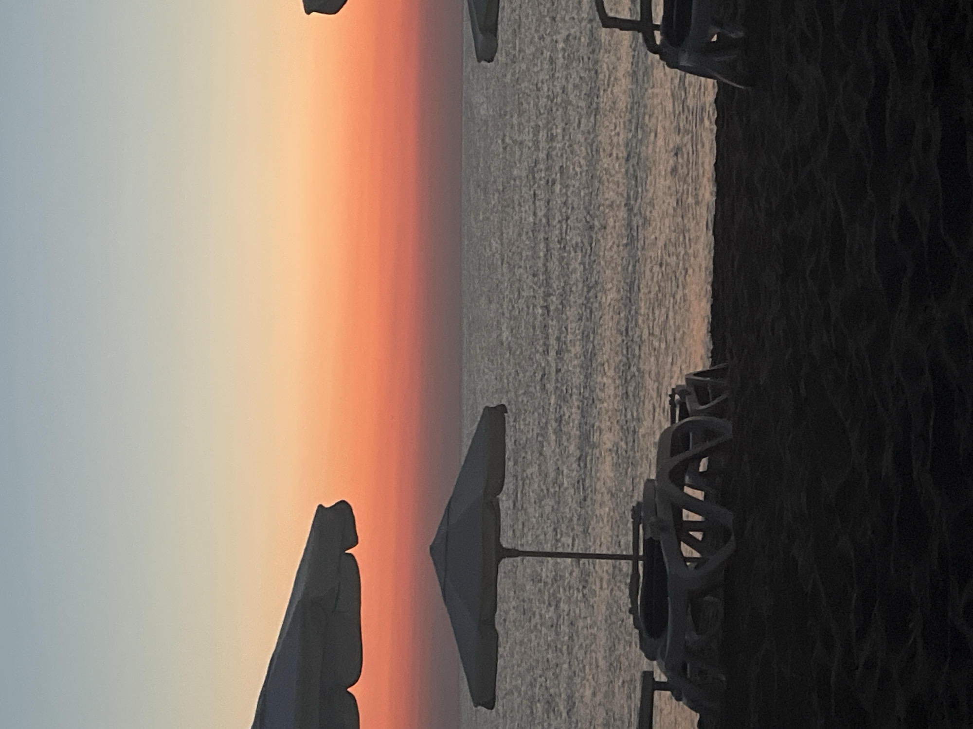 The image depicts a serene beach scene at sunset or sunrise in Lozenets, Bulgaria. The sky transitions from a pale blue at the top to vibrant hues of orange and pink near the horizon, casting a soft glow over the calm sea. In the foreground, dark sand covers the beach, with several empty white lounge chairs and umbrellas scattered across it, suggesting a quiet moment before or after a busy day. One large white umbrella is prominently featured in the middle ground, casting a shadow on the sand. Another umbrella is partially visible on the left side of the image, and a portion of a third umbrella is seen on the right. The water is a muted gray, reflecting the colors of the sky, and the gentle waves create a subtle texture on its surface. The overall atmosphere is peaceful and tranquil, characteristic of a seaside resort during off-peak hours.