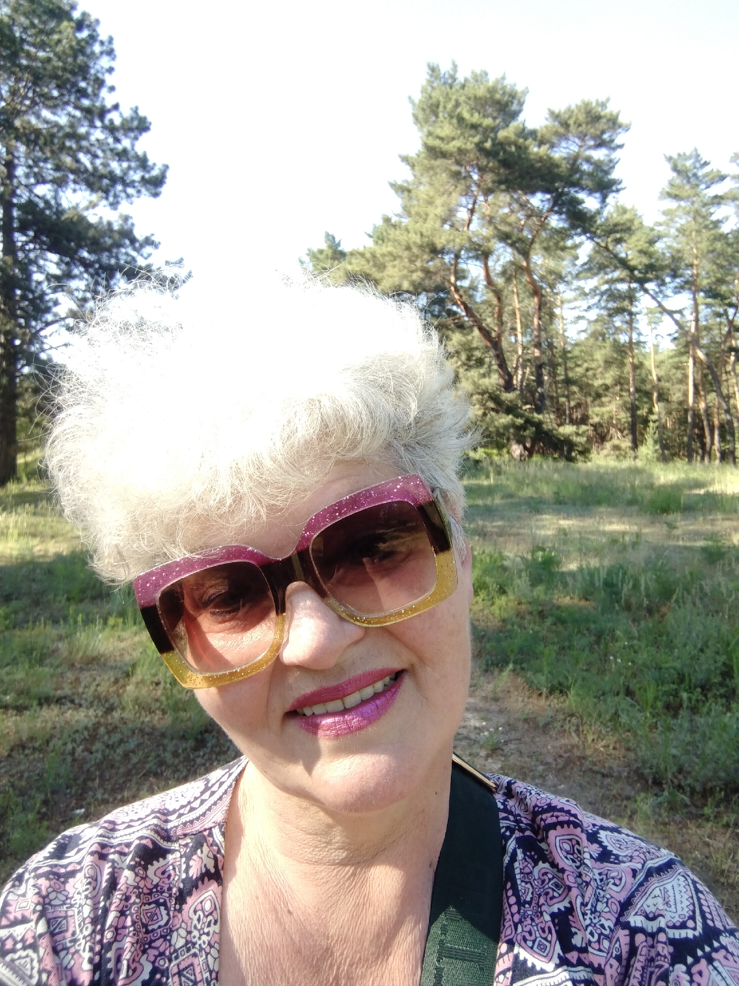 The image is a close-up selfie of an older adult woman, likely taken in Horishni Plavni, Ukraine. She has voluminous, bright white curly hair, which is illuminated by direct sunlight. She is wearing large, fashionable sunglasses with a glittery, two-toned frame, transitioning from dark pink/purple at the top to gold at the bottom. Her lips are painted with bright pink lipstick, and she is smiling. Her attire includes a patterned top in shades of purple, pink, and black.
The setting is outdoors in a natural environment, appearing to be a pine forest or woodland. Tall green pine trees are visible in the background, along with grassy ground. The bright lighting suggests it is daytime, under clear, sunny weather. The woman's smile conveys a cheerful or contented emotion. There is no visible text.