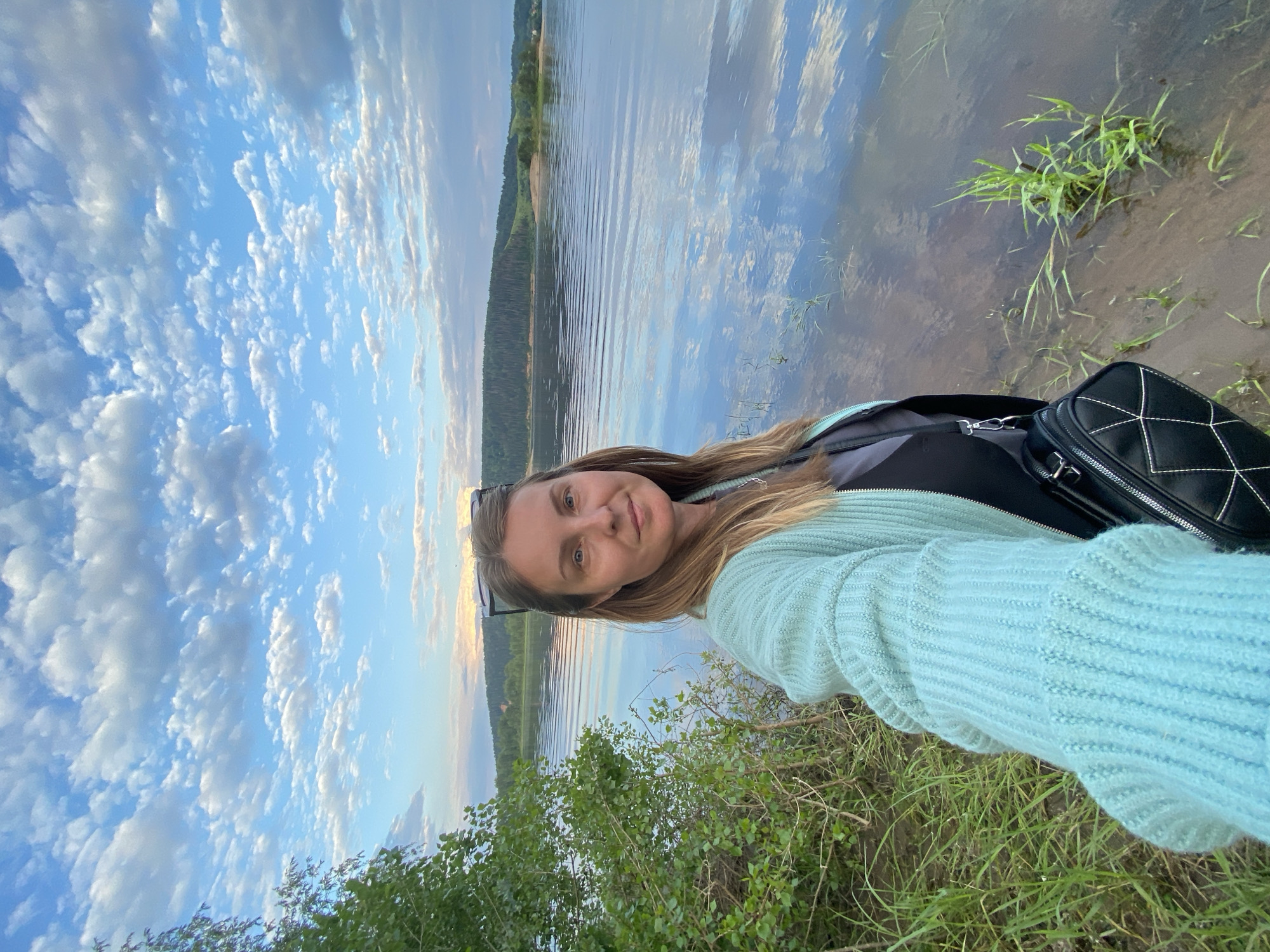 This media file is a portrait of a woman standing on the bank of a body of water, likely a river or lake, in Ольховка, Russia. The woman is facing the camera, taking a selfie, and smiling gently. She has long blonde hair and is wearing a light blue knitted sweater and a black backpack.
The scene is set at what appears to be either sunrise or sunset, given the soft lighting and the warm tones in the sky. The sky is filled with scattered white clouds against a bright blue background. The water reflects the sky and the distant treeline, creating a peaceful and serene atmosphere. The foreground shows grassy terrain and some green foliage.
The overall mood is relaxed and contemplative, suggesting the woman is enjoying a moment of solitude in nature. There is no visible text in the image.