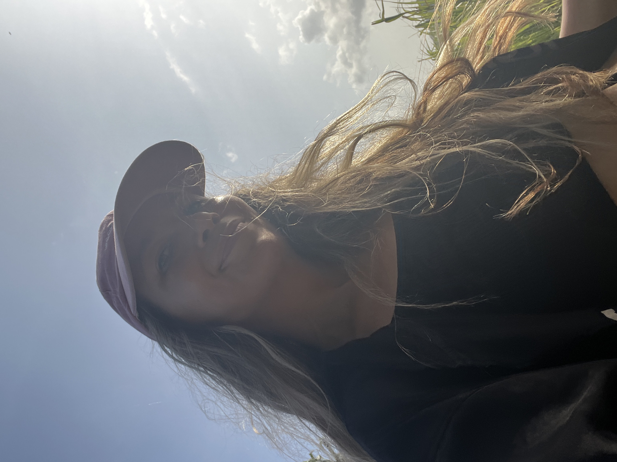 A low-angle selfie shows a young woman wearing a pink baseball cap and a black top. She has long, light brown hair that is being blown by the wind. The background is a bright blue sky with scattered white clouds. The sun is shining, creating a lens flare on the right side of the image. The woman is smiling and looking slightly off-camera. The overall impression is one of a casual, outdoor portrait. The location is Talalla, Sri Lanka.