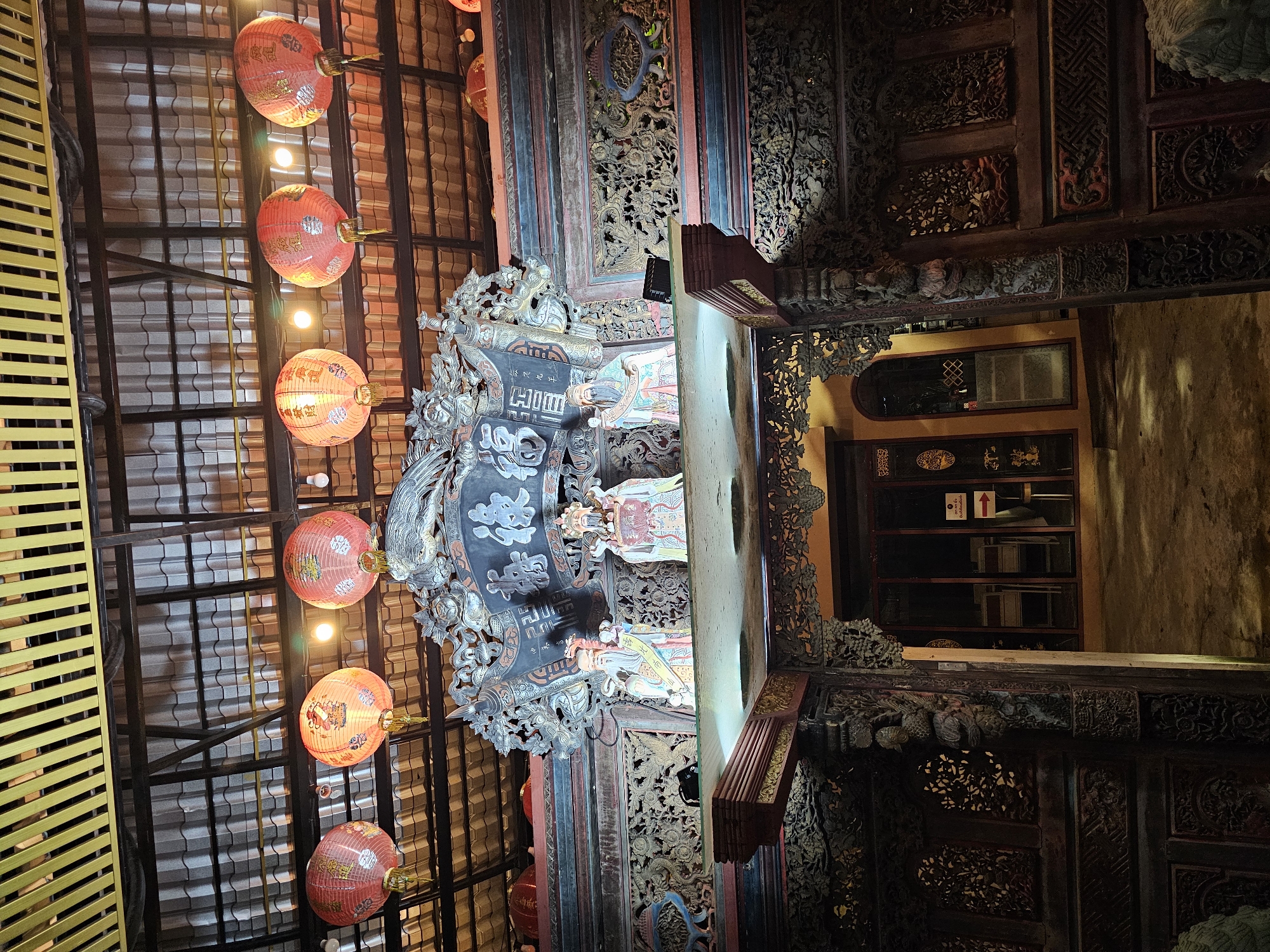 This image captures the ornate entrance of a traditional Chinese temple, likely located in Bangkok, Thailand, given its architectural style and common decorative elements found in the city's Chinese community areas such as Yaowarat (Chinatown).
The focal point is a highly decorated wooden archway with intricate carvings. Above the arch, a large, dark-colored plaque features Chinese calligraphy, flanked by carved mythical creatures, possibly dragons or phoenixes. Three figures, likely deities or historical figures from Chinese culture, are positioned on a platform above the archway. They are depicted in colorful traditional attire.
Dozens of red, round lanterns are suspended from the ceiling structure, casting a warm, ambient glow. These lanterns appear to be made of paper or silk and some have gold Chinese characters or imagery printed on them. The ceiling itself is constructed of interlocking wooden beams and corrugated roofing.
The entrance to the building behind the archway is visible, with glass doors and windows. The interior is dimly lit, with some furniture or seating visible. The ground in front of the entrance appears to be concrete or packed earth. The overall impression is one of cultural heritage and vibrant tradition, illuminated by the festive glow of the lanterns. The dark, carved wood and detailed figurines suggest an older, well-established place of worship or cultural center.