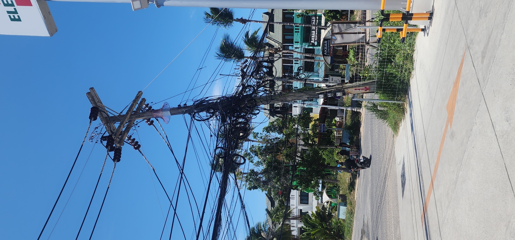 This image depicts a street scene in the city of Ko Samui District, Thailand, characterized by a tangle of electrical wires on a utility pole in the foreground. A motorcycle with two riders is visible on the road, moving away from the camera. Behind the motorcycle, several buildings with balconies and palm trees are present, suggesting a tropical or subtropical location. A sign for "AKBAR INDIAN RESTAURANT" is also visible on one of the buildings. The sky is clear blue, and the day appears sunny. The overall scene conveys a sense of everyday life in a bustling urban or semi-urban environment.