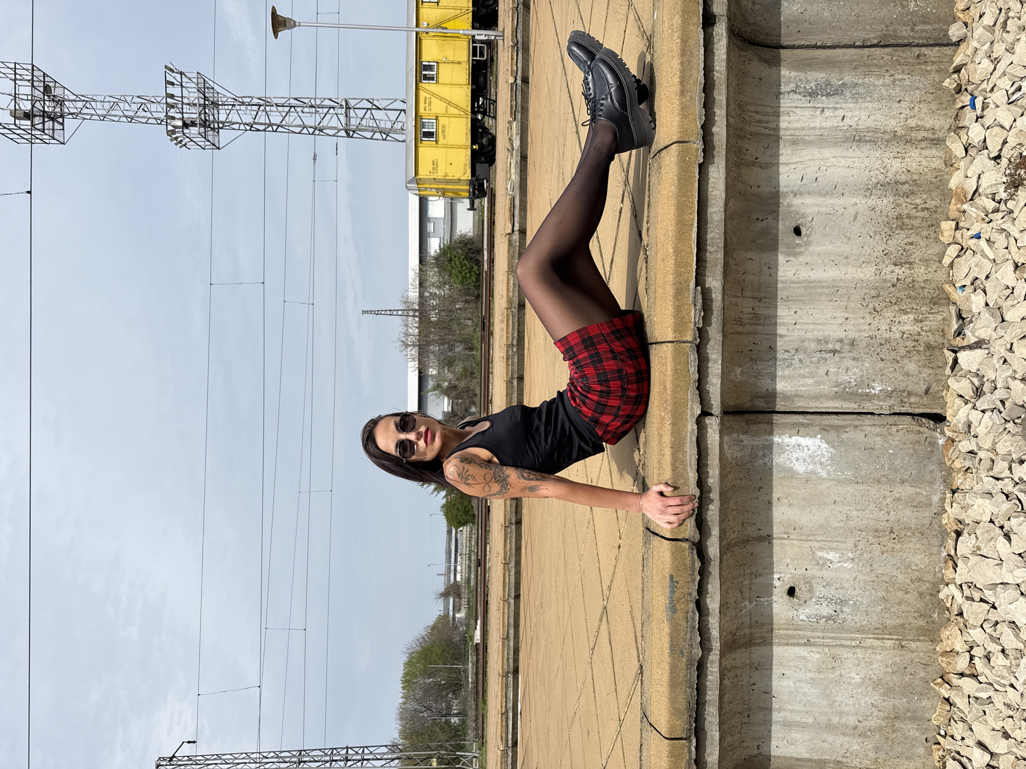 A woman is lying on her side on a concrete structure, likely a railroad platform edge. She is wearing a black tank top, a red and black plaid skirt, black tights, and black platform shoes. She has dark hair, sunglasses, and a visible tattoo on her arm.
The location appears to be a railroad area, with tracks visible in the background and a yellow railcar. There are overhead power lines and support structures. The ground is a combination of concrete, gravel, and some vegetation.
Given the mention of Pleven, Bulgaria, it's plausible that this scene is located in or around the Pleven railway station or a similar rail-related area in the city.