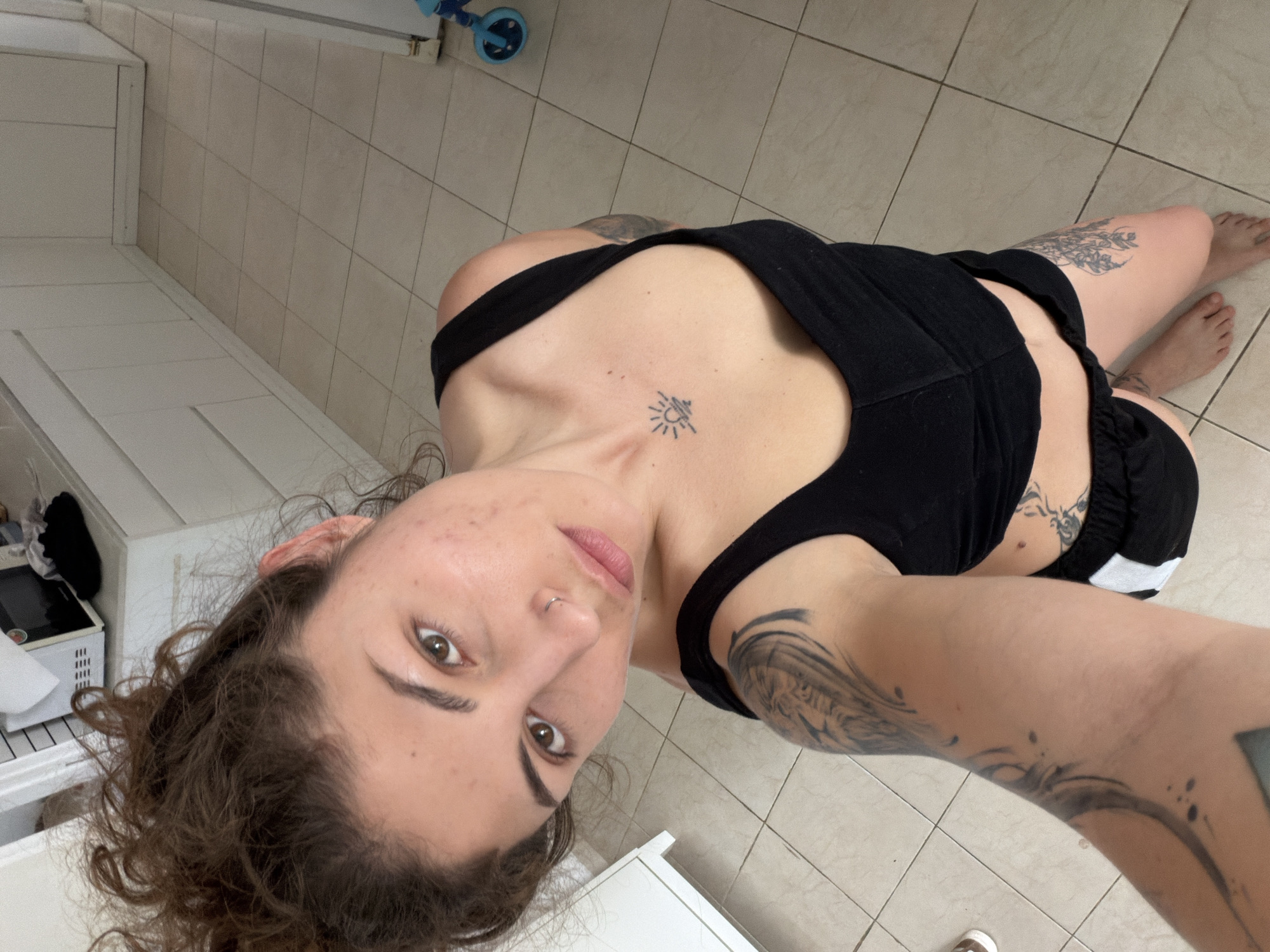 A woman is seen from a high-angle perspective indoors. She wears a black tank top and shorts, and she has several tattoos on her upper chest and arms. She looks directly at the camera with a neutral expression. The background consists of light-colored tile flooring and white cabinets/walls. A child's ride-on toy with blue wheels is partially visible in the upper background. The photo appears to be taken inside a residential location, likely in Kiryat Bialik, Israel, given that is the specified location.