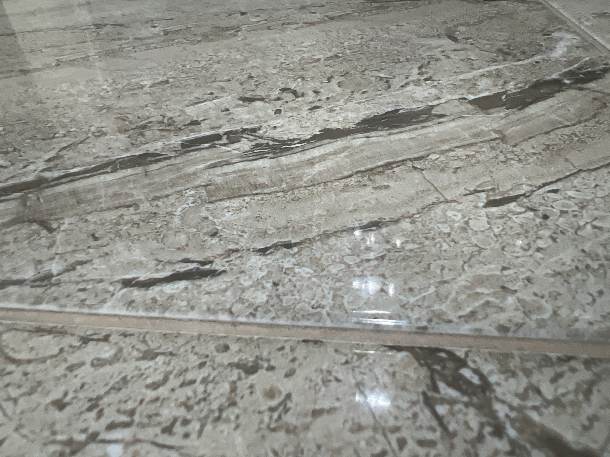The image displays a close-up, angled view of a tiled floor. The tiles have a marbled pattern with veins of brown, beige, and gray, mimicking natural stone. There are two distinct tiles visible, separated by a thin grout line running horizontally across the lower half of the image. The surface appears to be smooth and slightly reflective, with a few subtle highlights indicating overhead lighting. No people, objects, or actions are present in the frame. The environment is an indoor setting, likely a room with a tiled floor, possibly in a residential or commercial building. The specific location in Chernorechenskoye, Russia, cannot be determined from the visual information provided.