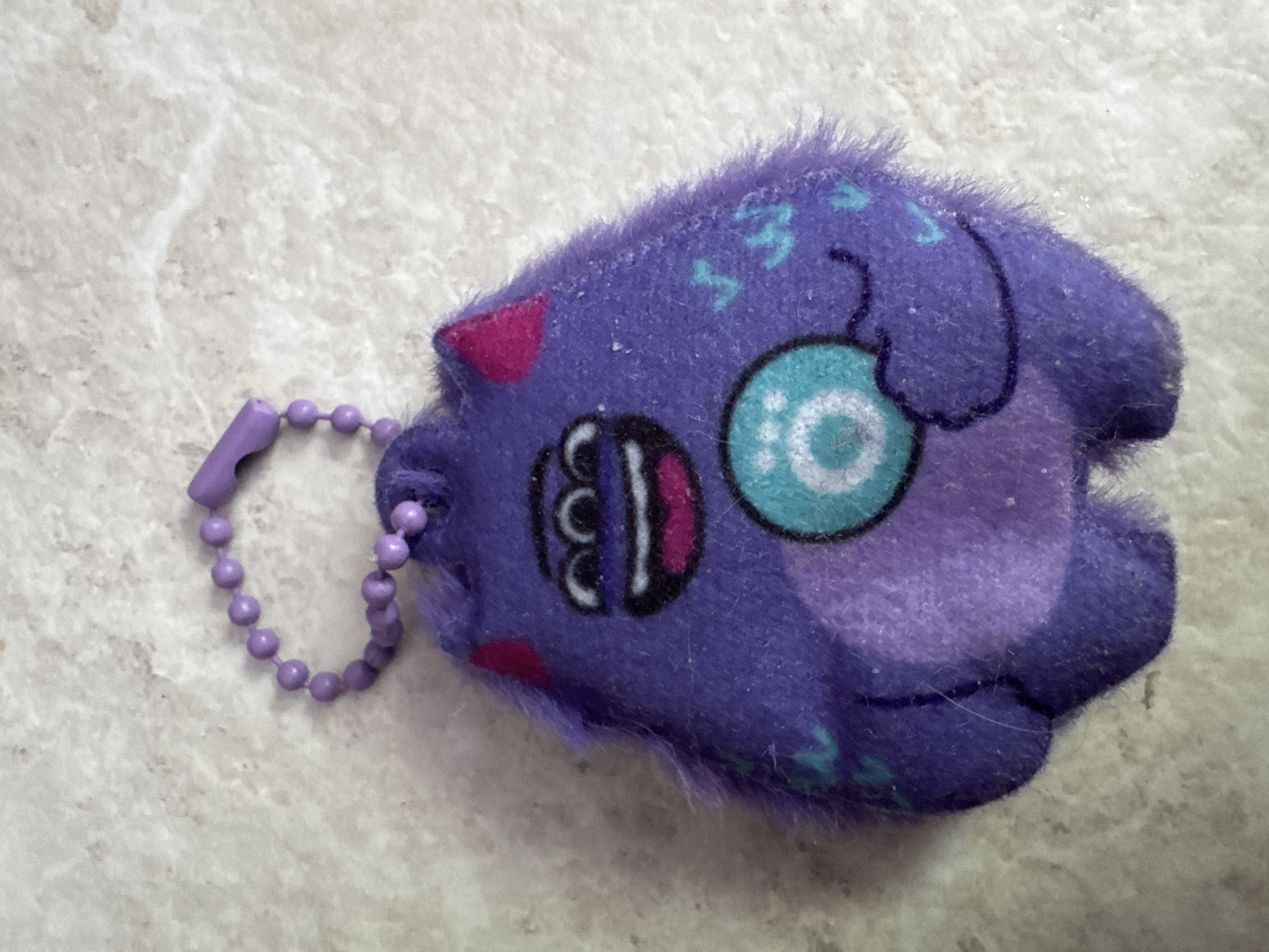 A purple plush keychain of a three-eyed monster with a teal orb in its hand is positioned on a light-colored, textured surface. The monster has pink triangle ears, a wide smile with a pink tongue and black lips, and a teal orb with a white spiral design. A thin purple beaded chain is attached to the top of the monster's head, leading to a purple plastic clasp. The background surface resembles a speckled countertop or tiled floor. There are no people or animals present in the image, and no identifiable landmarks or features that would suggest a specific location in Saratov, Russia, or any other city. The lighting appears to be overhead, casting a soft shadow to the left of the object.