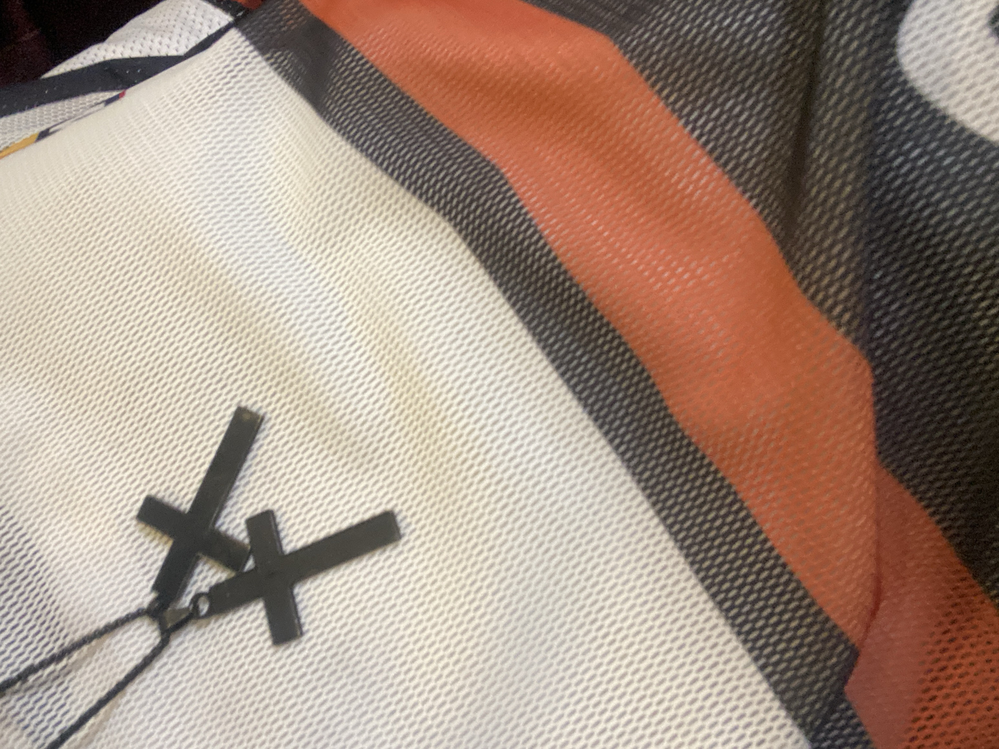 The image presents a close-up view of a textured fabric, possibly a garment or blanket, laid out flat. The fabric features a striped pattern composed of white, dark grey, and muted orange-red sections. The white and orange-red areas exhibit a distinct mesh or breathable weave, characterized by small, repeating patterns of diamond-shaped holes. The dark grey stripes appear to have a denser, diagonal woven texture.
Resting on the white section of the fabric are two small, black, cross-shaped charms. These crosses are connected by small rings to a thin, black cord, suggesting they are part of a necklace, bracelet, or similar accessory. One cross is slightly elevated and positioned above the other, causing a slight overlap. The charms cast soft shadows on the fabric, indicating diffused lighting. No people or specific activities are depicted. There is no visible text. The setting appears to be an indoor environment. The provided location context for this media is Badagry, Nigeria, though no specific visual cues within the image confirm this geographical location.