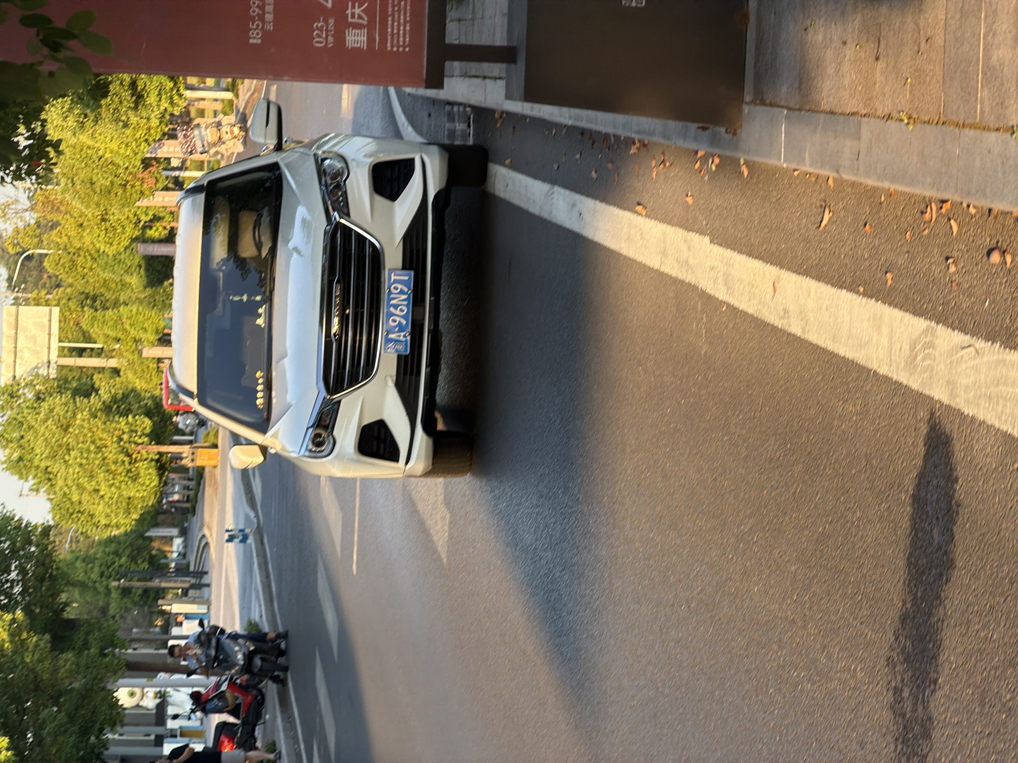 A white SUV is parked on a city street in Jiangjin, China. The vehicle, with license plate A-96N9T, is positioned at an angle to the right, facing forward. The street is paved with asphalt and marked with a white line. To the right of the SUV, a sidewalk with paving stones and a red sign is visible. The sign has Chinese characters and numbers, including "185-99" and "重庆" (Chongqing). Trees with green foliage line the street, and in the background, more trees and buildings can be seen. Sunlight casts shadows on the road, indicating it is likely late afternoon. There are no people in the immediate vicinity of the SUV.