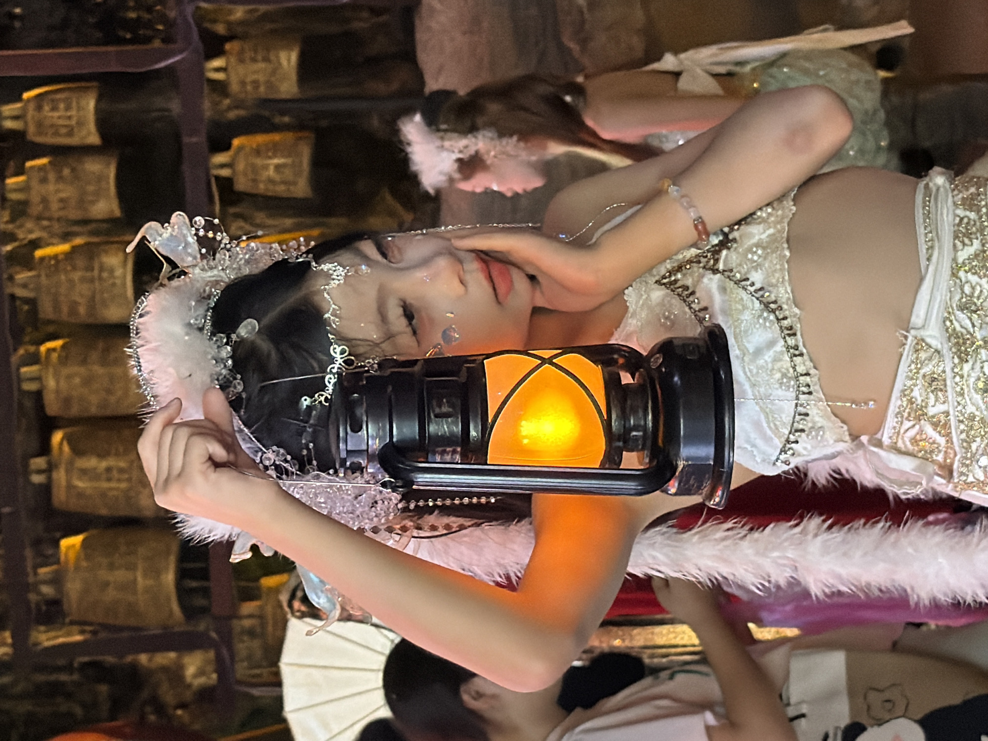 In the Yuzhong District of China, a young girl, adorned with a white feather and crystal headpiece, poses with a lit black lantern in front of a backdrop of decorative bells. She wears a white, ornate costume with gold embellishments and a feathery white boa. Her right hand rests on her cheek, and she is looking to the side. In the background, another young woman, also in costume, is visible. The scene appears to be an indoor event or performance.