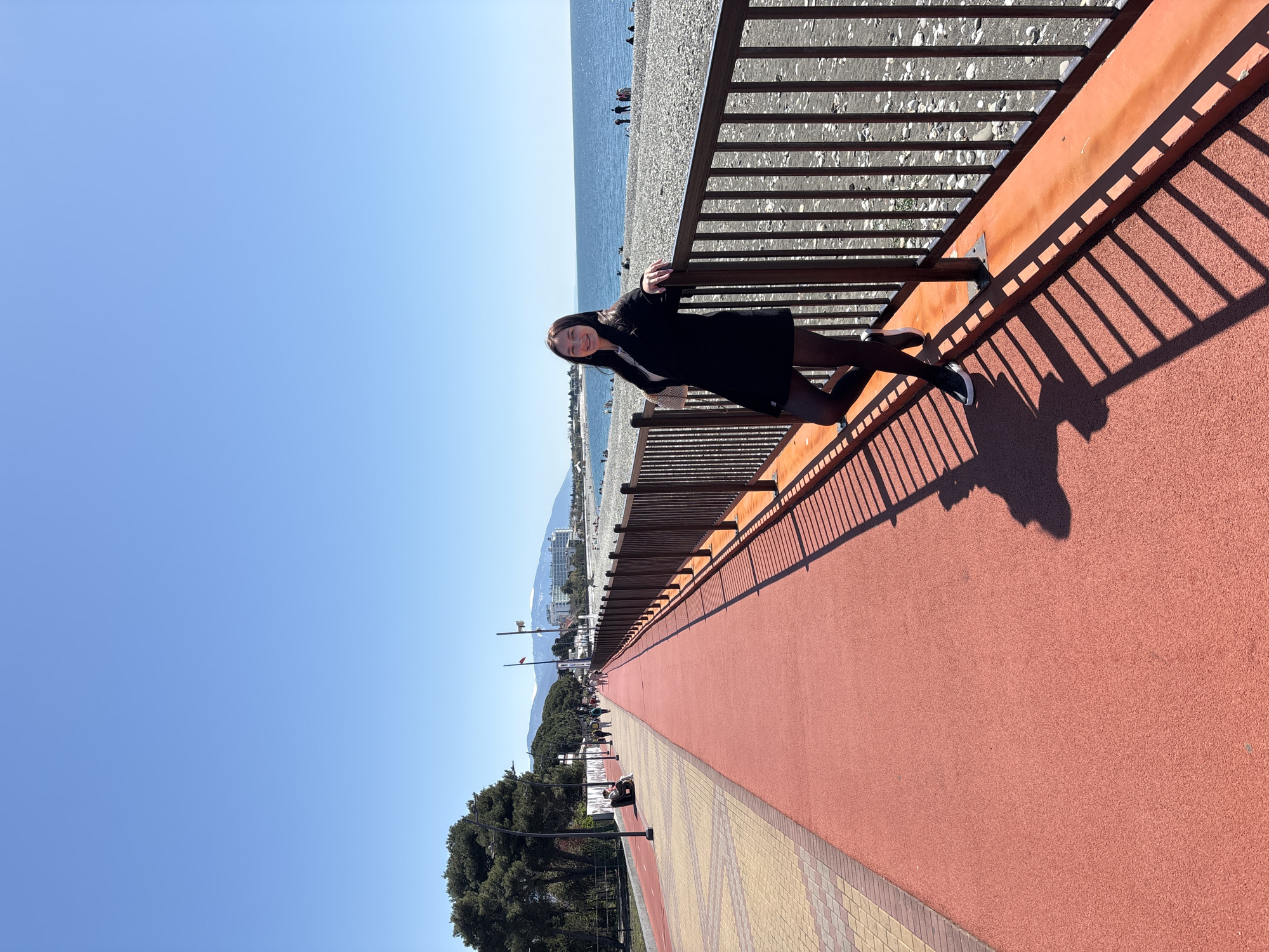 This image presents a bright and sunny coastal scene in Sochi, Russia, taken from an elevated perspective. The central subject is a young woman with dark hair, wearing a black coat or dress, dark leggings, and light-colored sneakers. She is standing on a wide, reddish, textured track or path, leaning against a dark brown wooden railing, and smiling directly at the camera.
The railing extends horizontally across the image, separating the track from a broad, pebbly beach and the calm, blue sea. In the distance, along the coastline, various buildings are visible, backed by majestic mountains, some of which appear to have snow-capped peaks. The sky is clear and a vibrant blue, suggesting excellent weather conditions.
Strong, direct sunlight casts long, diagonal shadows from the railing onto the reddish track, indicating either a late morning or early afternoon time. To the left of the main track, a section of light brown and yellow checkered pavement is visible, running parallel to the track. Further inland, a line of green trees and lampposts extends into the distance. Several other people are scattered along the pebbly beach and further down the promenade, enjoying the day. The overall impression is one of leisure and relaxation in a beautiful, sunny environment. There is no visible text in the image.