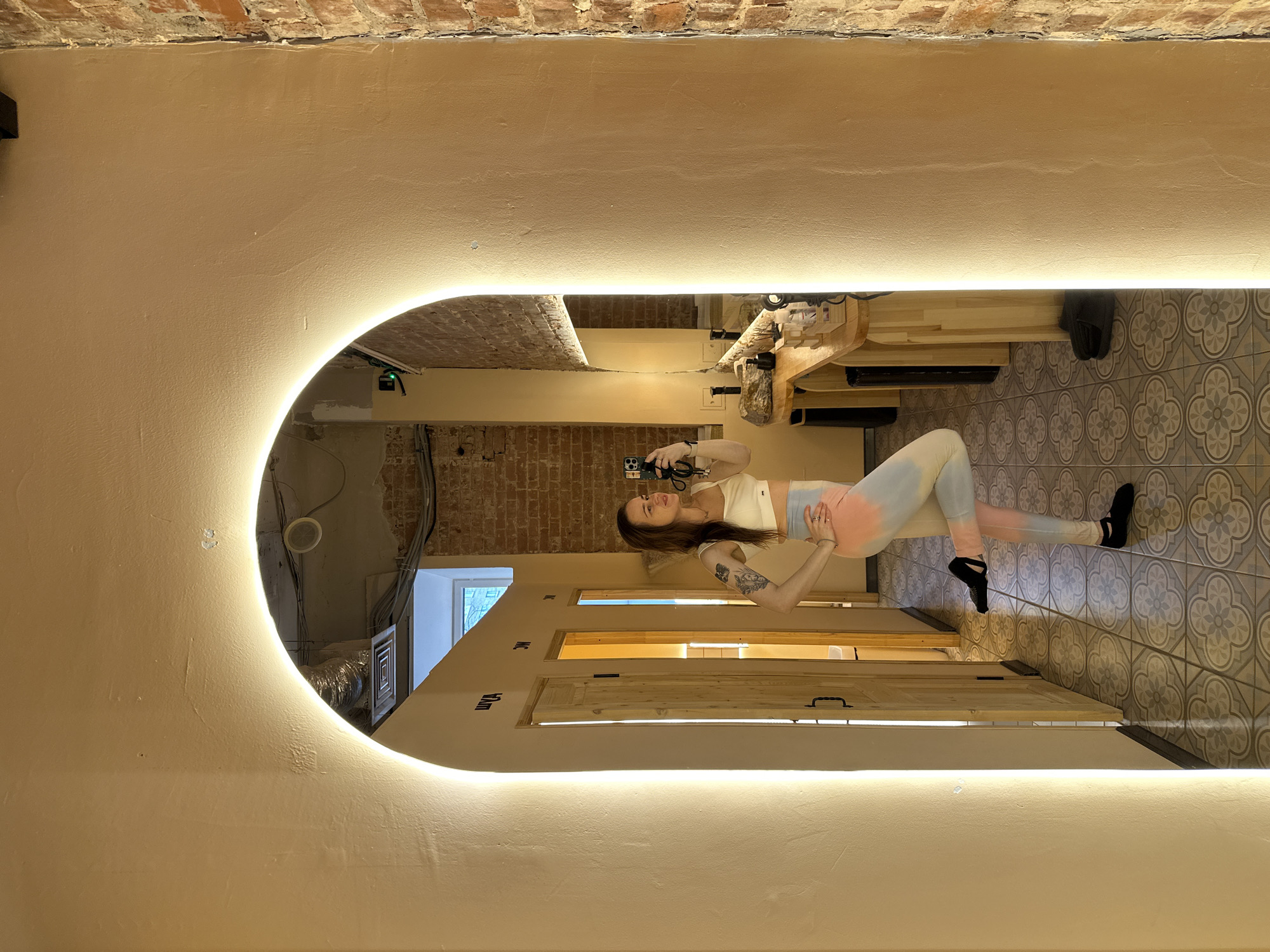 A mirror reflects a woman holding a smartphone, presumably taking a photo of herself in a hallway. The location is suggested to be in Moscow, Russia, but the context lacks visual clues to verify that. She is wearing workout attire, including a white top and tie-dye leggings. The hallway has doors on either side, leading to other rooms. The floor has a gray and white patterned tile, and the walls are painted a light beige. The mirror is long, rectangular, and rounded on one end, with integrated lighting. The ceiling appears to be partially exposed brickwork. In the reflection, a room with a wooden countertop and various items is visible.