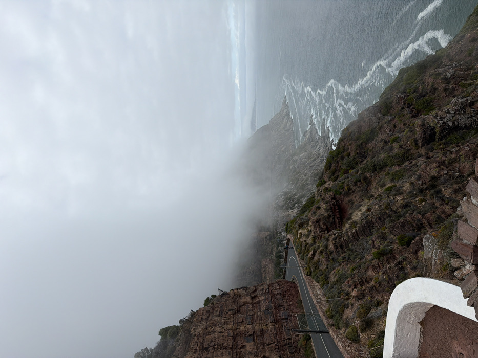 The image displays a coastal scene on Chapman's Peak Drive, located near Hout Bay in Cape Town, South Africa. The environment is overcast and foggy, with a uniform grey sky. On the left, a steep, reddish-brown rock cliff face rises vertically, featuring visible geological strata and sparse dark green vegetation. Protective netting is affixed to sections of the upper cliff. A two-lane asphalt road, distinguished by a low stone barrier along its seaward edge, curves along the base of the cliff and extends into dense fog in the mid-ground. To the right of the road, vegetated slopes descend towards the ocean. The dark blue-grey Atlantic Ocean occupies the right side of the frame, exhibiting white surf and foam along the rocky shoreline. Distant coastal features are largely obscured by the fog. In the bottom-left foreground, a white, curved concrete wall and reddish-brown paved ground are visible, suggesting a viewing area. No individuals, vehicles, or specific actions are present in the scene.