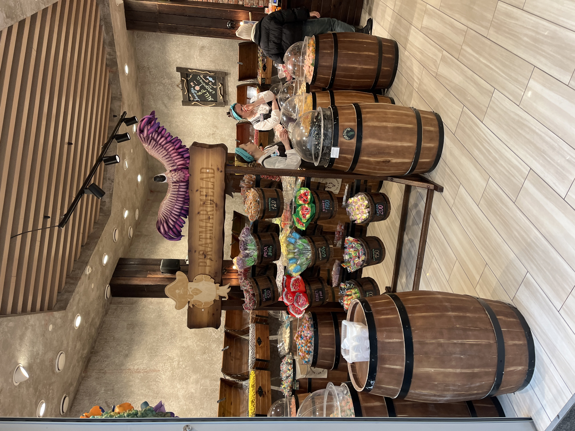 Here is a description of the image:
The image captures the interior of a candy store called "PIRATMARMELAD" in Krasnoyarsk, Russia. The store's design incorporates a nautical theme, with displays resembling wooden barrels filled with various colorful candies. Customers, some dressed in period costumes, are seen examining the candies. There are price tags on some of the candy barrels. The lighting is bright, illuminating the candies and interior design.