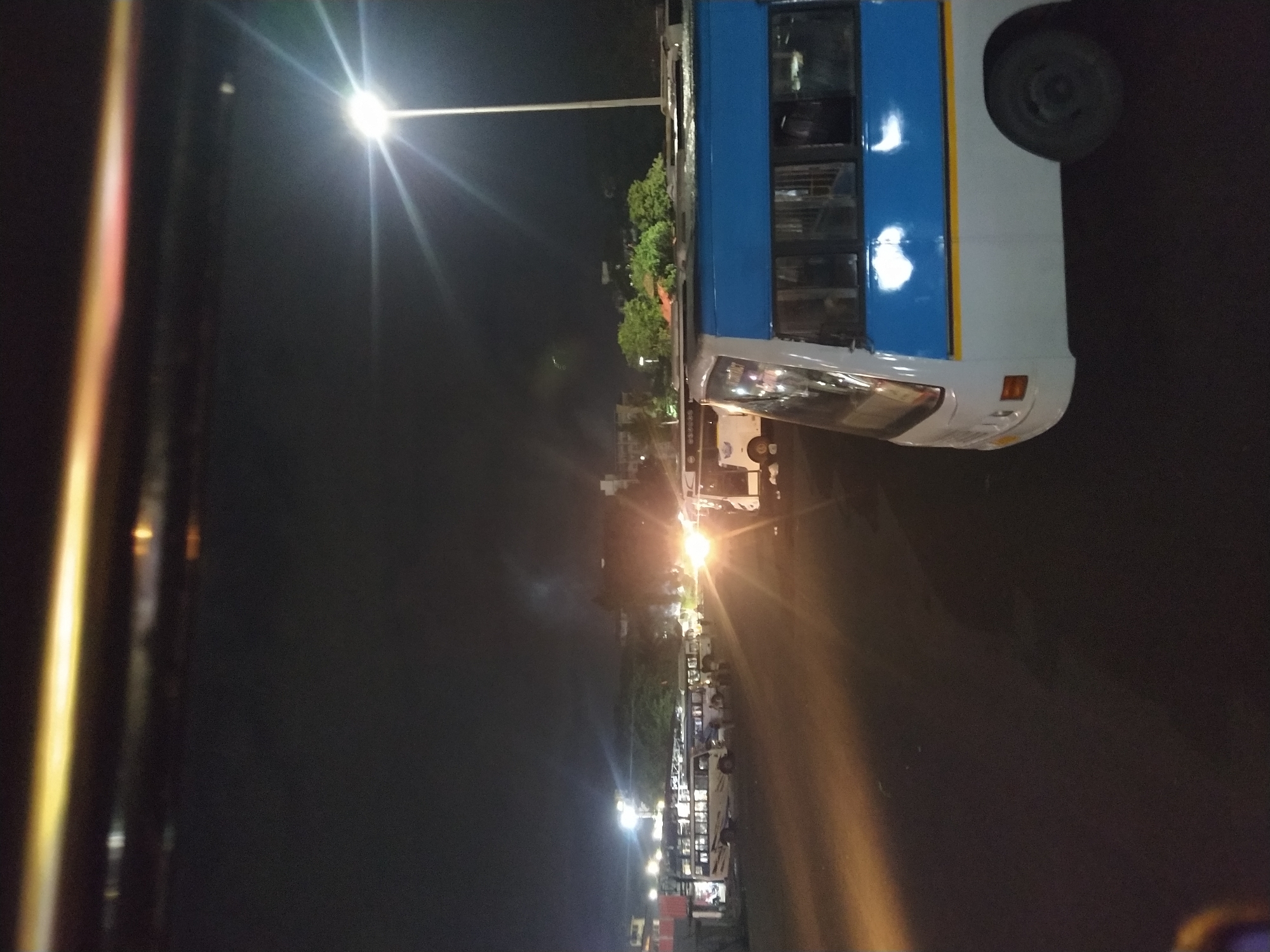 The image depicts a night scene in Margao, India, showing a bus station with several buses parked under streetlights. The focus is on a white and blue bus in the foreground. A street light is visible above the bus. The surrounding environment shows the silhouette of trees, buildings, and parked cars or buses. The overall scene gives the impression of a quiet bus station at night.