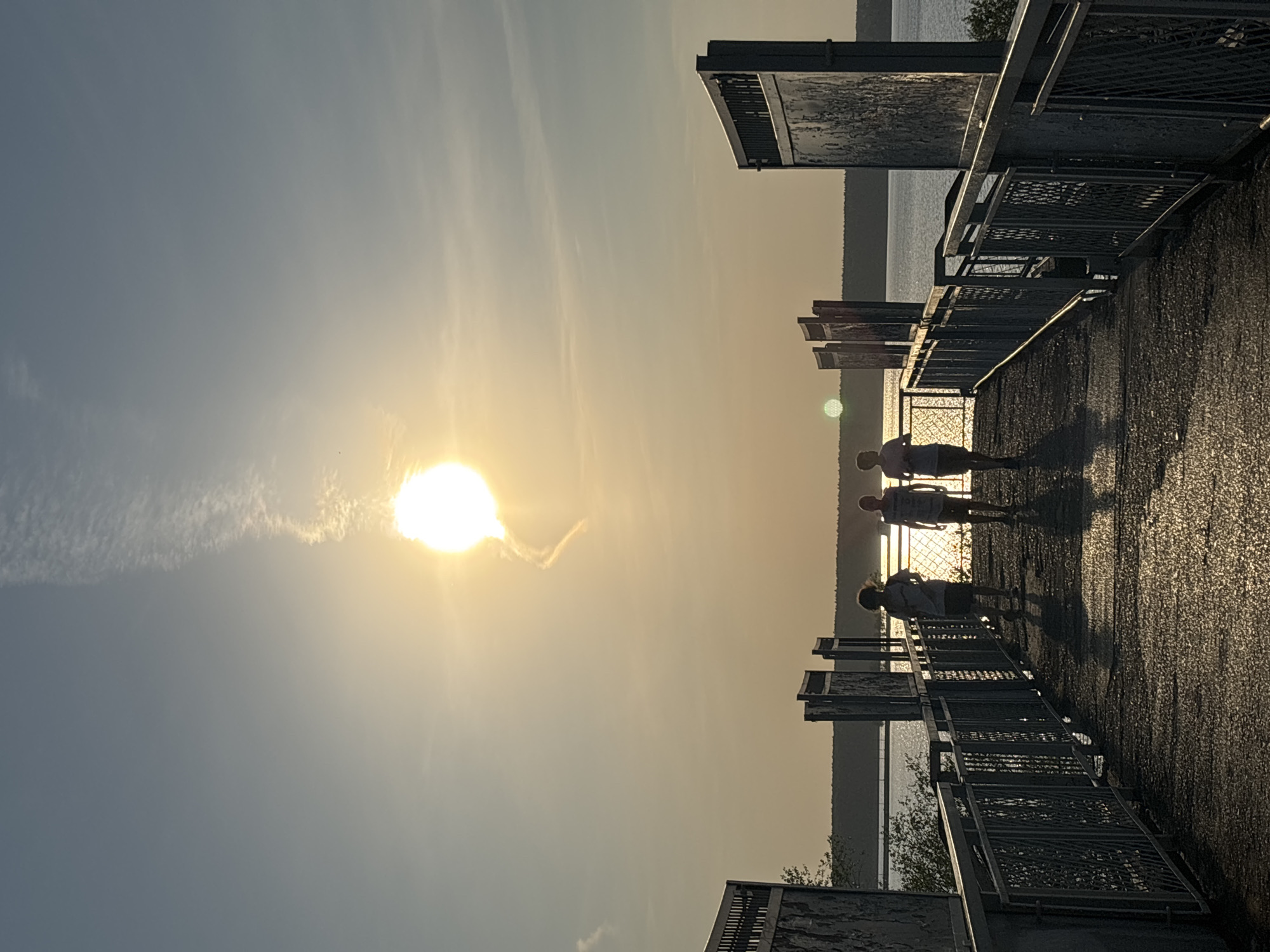 The image captures a scene at sunset on a pier in Perm, Russia. The sun is positioned prominently in the sky, causing a significant glare and long shadows to be cast on the pier's surface. The silhouettes of three people walking along the pier are visible, their forms obscured by the light. The pier has metal railings and platforms, and a body of water stretches out beyond the pier's edge, with land visible in the distance. The sky is a light blue fading into yellow as the sun gets closer to the horizon.