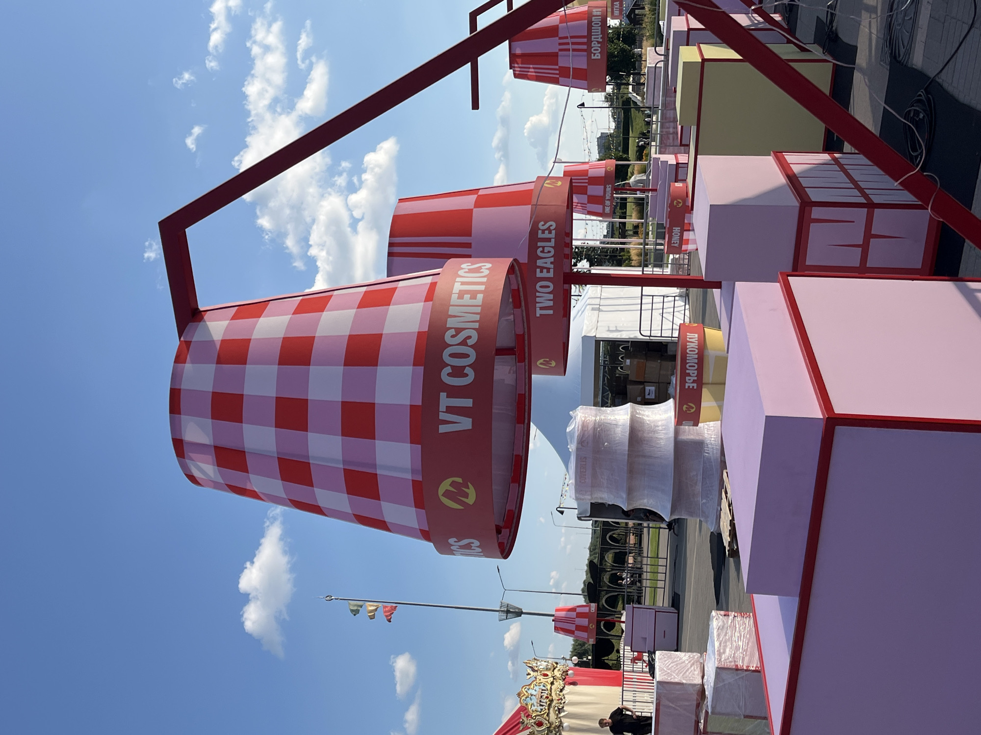 The image depicts an outdoor event space in Nizhny Novgorod, Russia, under a clear blue sky with scattered clouds. The foreground features several large, pink, cube-shaped structures with red trim, some stacked on top of each other. One cube has red lettering that reads "ЛУКОМОРе" (Lukomorye) upside down. Behind these structures, a large, red, frame-like structure extends upwards, supporting oversized, red and white checkered lampshade-like objects. These "lampshades" have red bands around their bases with text. One prominent band reads "VT COSMETICS" with a yellow logo. Another visible band reads "TWO EAGLES". Further back, other similar lampshades are visible, one with Russian text that appears to be "БОРДШОН #1" (Bordshon #1).
To the left, a brightly colored carousel with gold embellishments is partially visible, and a person in a dark shirt stands nearby. Tents and other structures are set up in the mid-ground. In the background, a tall pole with three small flags is visible, along with streetlights and a fence line. People are scattered throughout the background, suggesting a public gathering. The overall scene suggests an outdoor festival or promotional event.