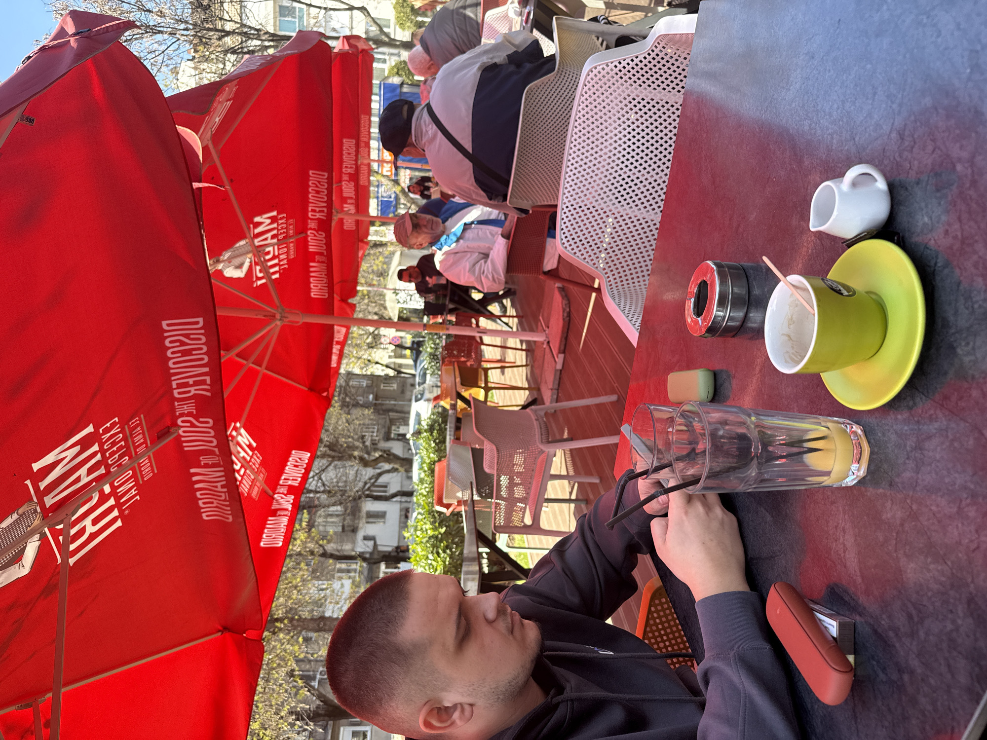 The image depicts an outdoor cafe scene in Plovdiv, Bulgaria. A man sits at a dark-colored table under a large red "WAM" branded umbrella. The table holds a glass of liquid, a green cup and saucer, a small white creamer, an ashtray, and a red cigarette case. The man is wearing a dark hoodie. Other patrons are seated in the background at tables with white perforated plastic chairs, with red umbrellas. Buildings and trees are visible in the background.