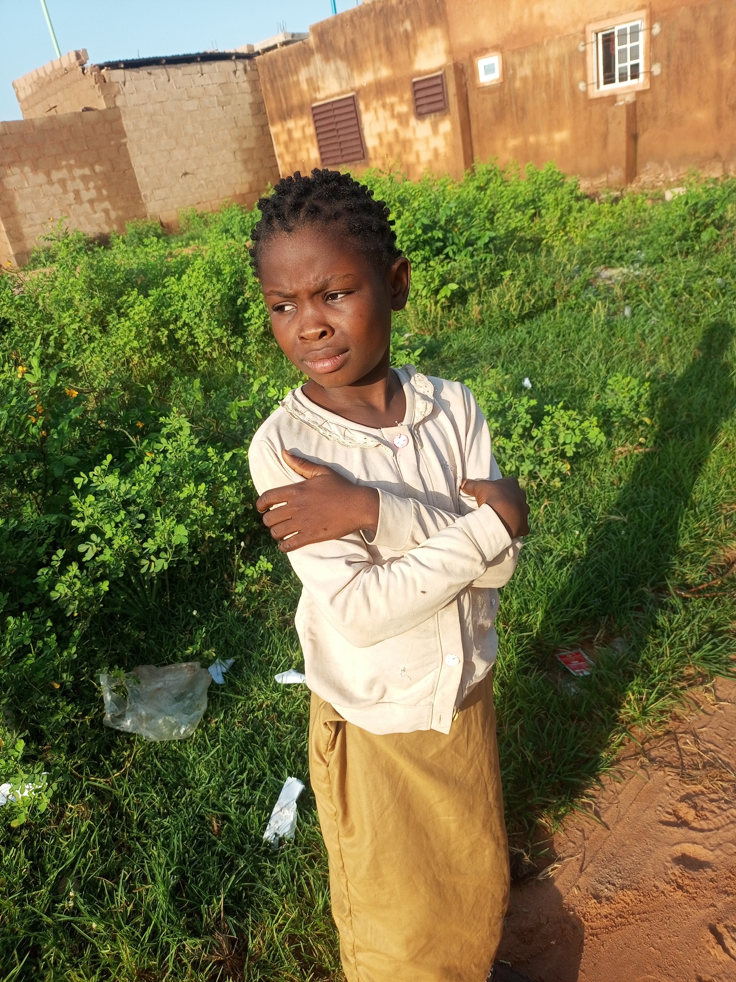 A young girl stands in a grassy area, looking to her left with her arms crossed. She is wearing a light-colored long-sleeved shirt and a tan skirt. Behind her, there is a brown building with windows. The ground is a mix of grass and dirt. There is some trash scattered on the ground. The lighting suggests it is either early morning or late afternoon.