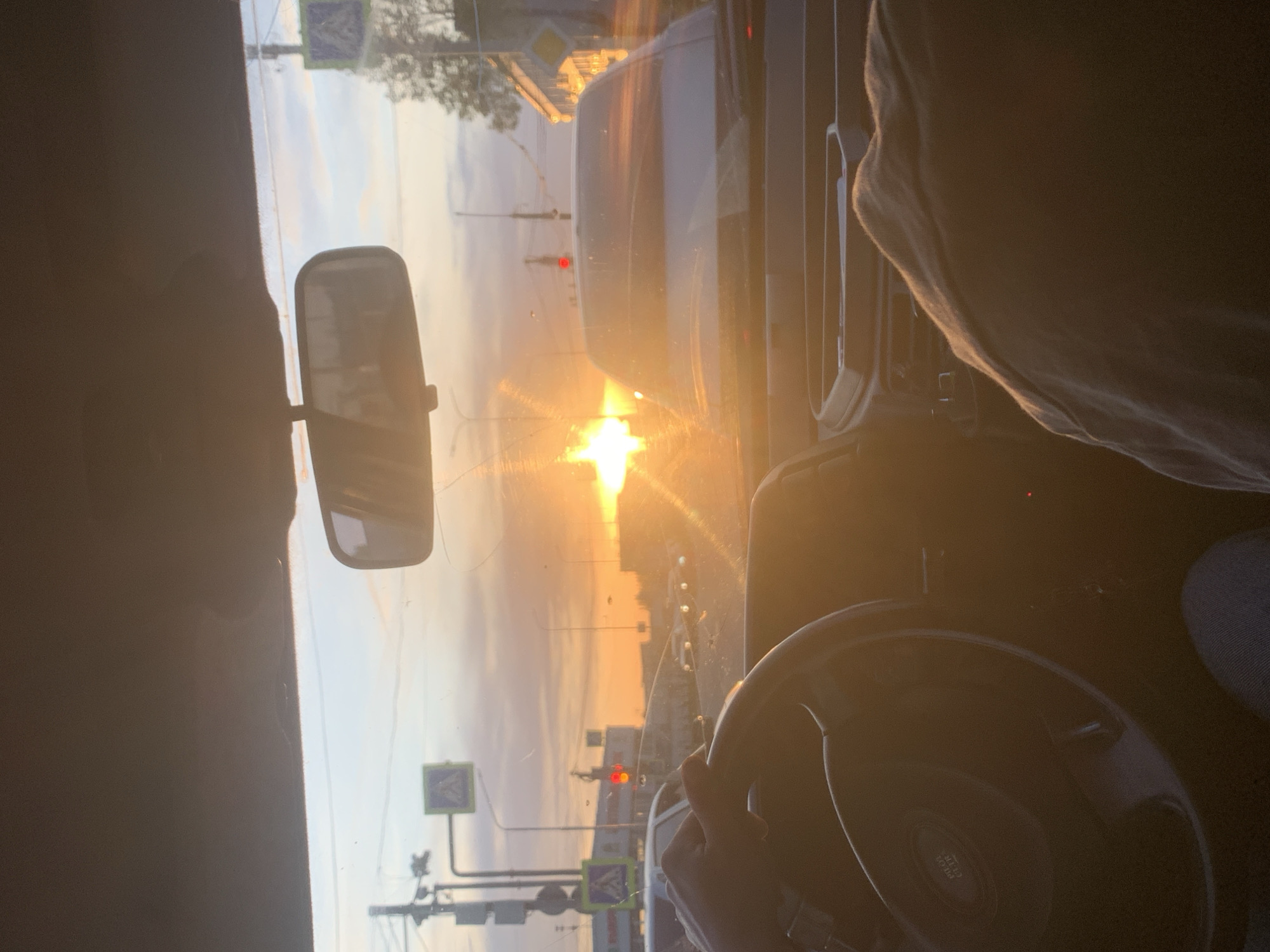 The photograph, taken from the interior of a vehicle, captures a view through the windshield at sunset in what is identified as the city of Chaikovsky, Russia. A rear-view mirror is visible in the upper-left quadrant of the frame. The sun, low on the horizon, casts a bright glare on the windshield. In the foreground, a steering wheel is partially visible, with a hand gripping it. Through the windshield, urban elements such as streetlights and road signs are visible. The sun's position and glare make it difficult to identify the precise location within Chaikovsky or specific landmarks. A vehicle is present in front of the vehicle taking the image. The overall setting is indicative of urban traffic at dusk.