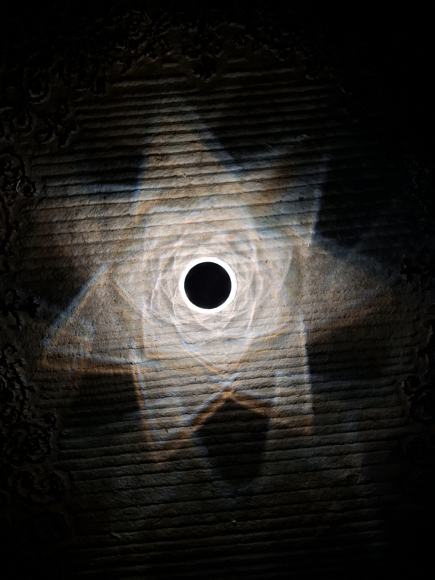 The image shows a close-up, overhead view of a textured surface, possibly a carpet or fabric, illuminated by a light source creating a circular halo of bright light in the center. Within the halo is a solid black circle, suggesting a lens or an object blocking light. The light casts geometric patterns and shadows across the textured surface, with faint prismatic colors visible in some areas. The edges of the image fade into darkness, with some textured detail visible on the far left and right, resembling a decorative border or distressed material. The scene does not contain any people, animals, or recognizable landmarks. Therefore, it is not possible to determine a specific location within Abu Dhabi, United Arab Emirates, or any other geographical context.