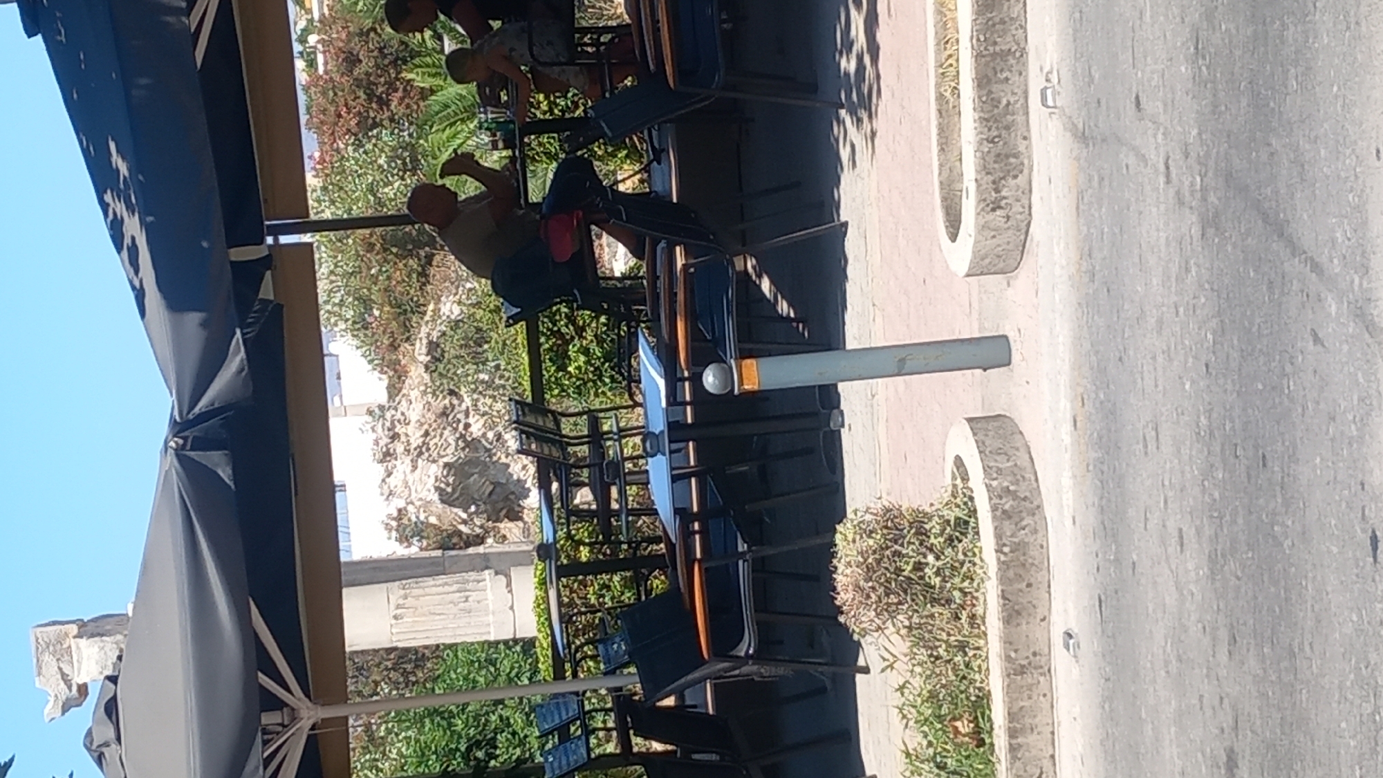 The image depicts an outdoor seating area of a cafe on a sunny day, likely in the city of Kos, Greece, given the architectural elements and vegetation. In the foreground, the pavement of a road is visible, with a concrete planter box containing dry foliage to the left, and another similar planter box to the right. A gray metal pole with an orange top stands vertically in the middle ground, positioned between the road and the cafe seating.
The cafe area itself is covered by a large black umbrella, providing shade. Several tables and chairs are arranged under the umbrella. Some tables have black surfaces with blue accents, and metal chairs with wooden seats and backs are visible.
In the background, a man and a child are seated at one of the tables. The man, wearing a light-colored shirt and dark pants, is leaning forward, appearing to interact with something on the table, possibly food or drink. The child, wearing a light-colored dress, is also seated at the table. Another person, partially visible, is seated at an adjacent table to the right, also appearing to be at the cafe.
Behind the cafe seating, there is a dense area of greenery, including trees with reddish leaves and palm-like plants. Further in the background, there are remnants of ancient stone structures, possibly ruins, with a clear blue sky overhead. The overall impression is a typical scene of outdoor dining or relaxation at a cafe in a location that combines modern amenities with historical surroundings.