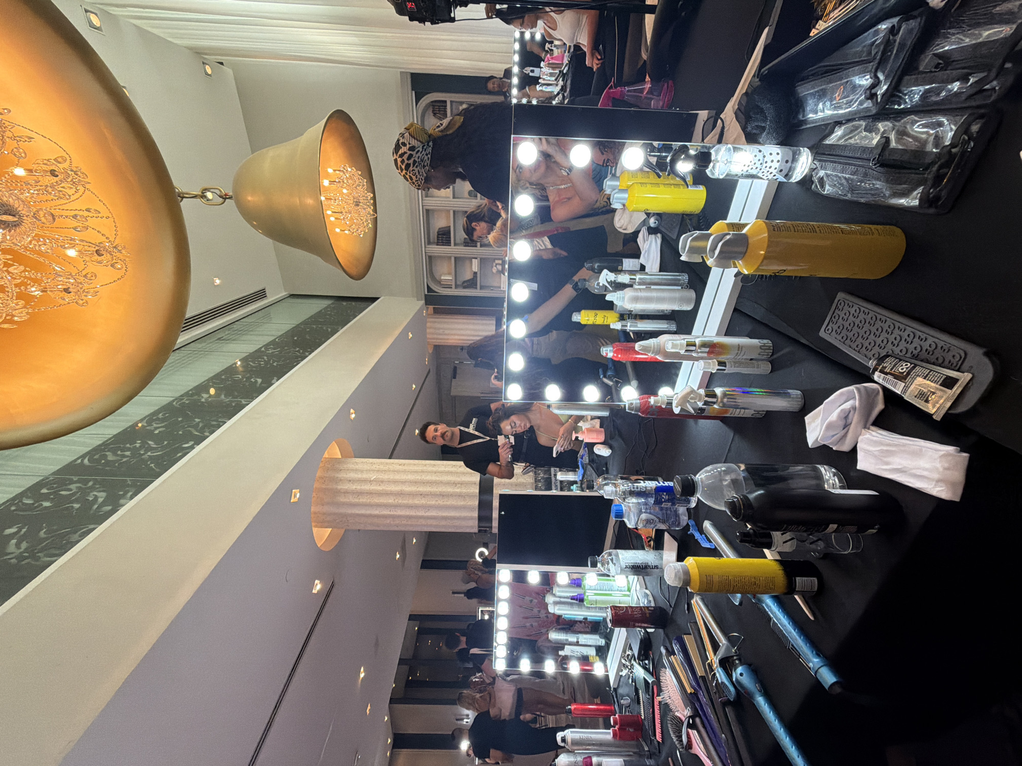 The image shows a bustling hair and makeup room, likely backstage at an event or fashion show in Miami Beach, United States of America. Several people are visible, including stylists attending to models or clients in front of lighted makeup mirrors. The surfaces are cluttered with hair and makeup products, tools, and accessories. A large, ornate chandelier is partially visible in the upper-left corner, indicating a luxurious setting. There are multiple hair and makeup stations set up, suggesting a high volume of activity. The atmosphere appears energetic and focused.