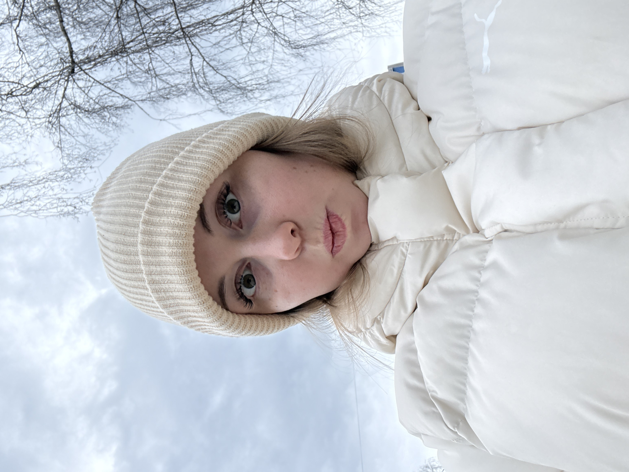 A young woman, possibly white, is wearing a light beige knitted hat and a light beige puffy jacket with a white Puma logo, gazing upwards. The background suggests an outdoor setting in winter, with a cloudy sky and bare tree branches visible.