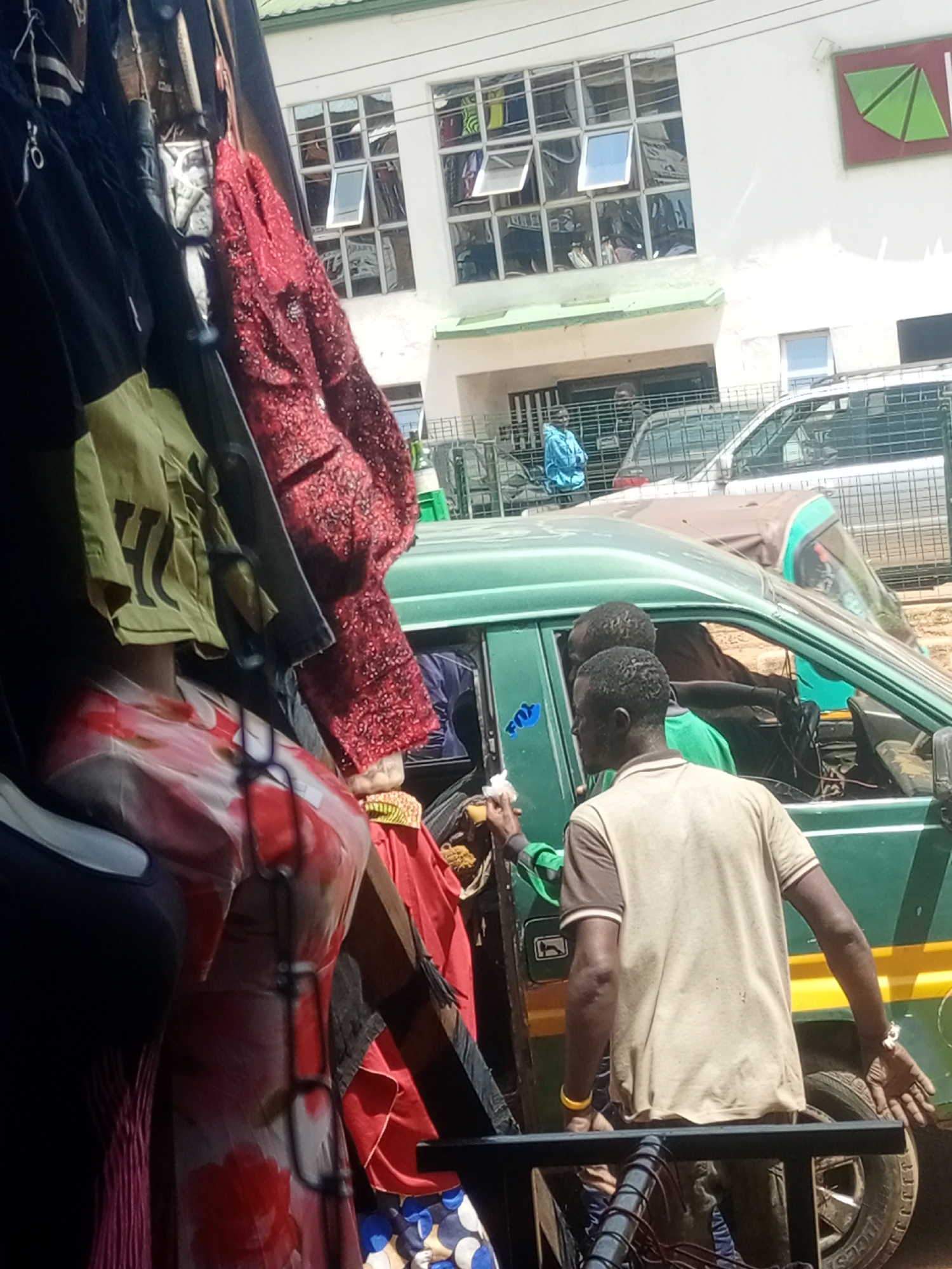 In Bukuru, Nigeria, a street scene unfolds with a focus on a vibrant marketplace and its surroundings. Foreground elements include a display of clothing on racks, featuring a red sequined jacket, a pale yellow t-shirt with black lettering, and a floral-patterned dress on a mannequin. Behind this, a green minibusses dominates the mid-ground, its side door open. Two men are visible within and near the bus. One man, wearing a tan t-shirt, stands by the open door, his right arm extended. Another man, seen through the open door, wears a green shirt. The bus is parked on a street, with other vehicles and structures visible in the background. A building with large, reflective windows and a green awning is in the upper background, suggesting a commercial area. The overall impression is of a busy urban environment.
