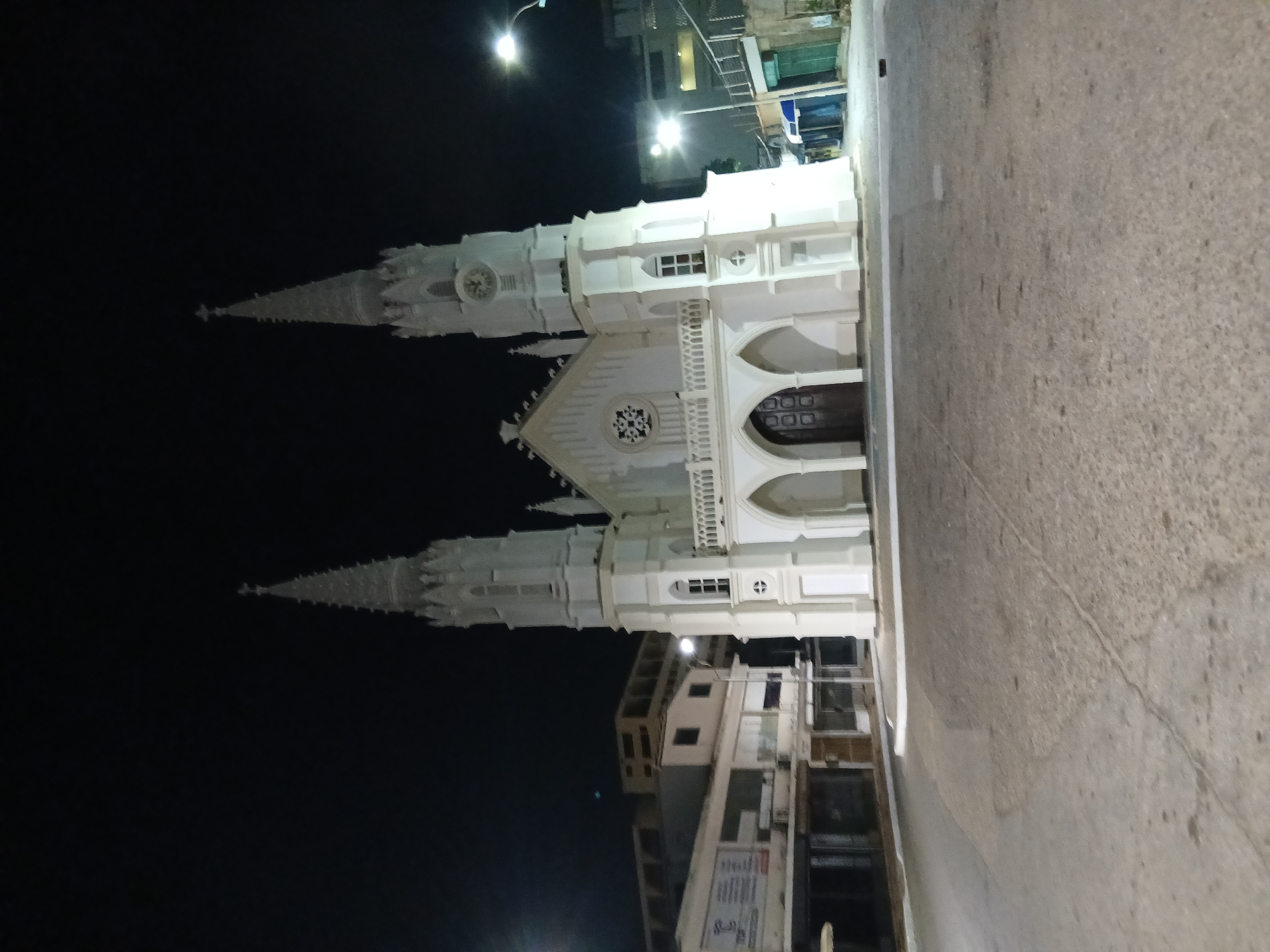 The image depicts the Iglesia San Juan Evangelista, a white, neo-Gothic church in Juan Griego, Venezuela, at night. The church features twin spires, a central rose window, arched doorways, and decorative elements, all illuminated. A street runs along the right side of the frame, showing a sidewalk and roadway surface. The sky is dark and clear, while buildings line the street behind and to the side of the church. A few streetlights are visible. The image is taken from a ground-level perspective, showing the full height of the church.
