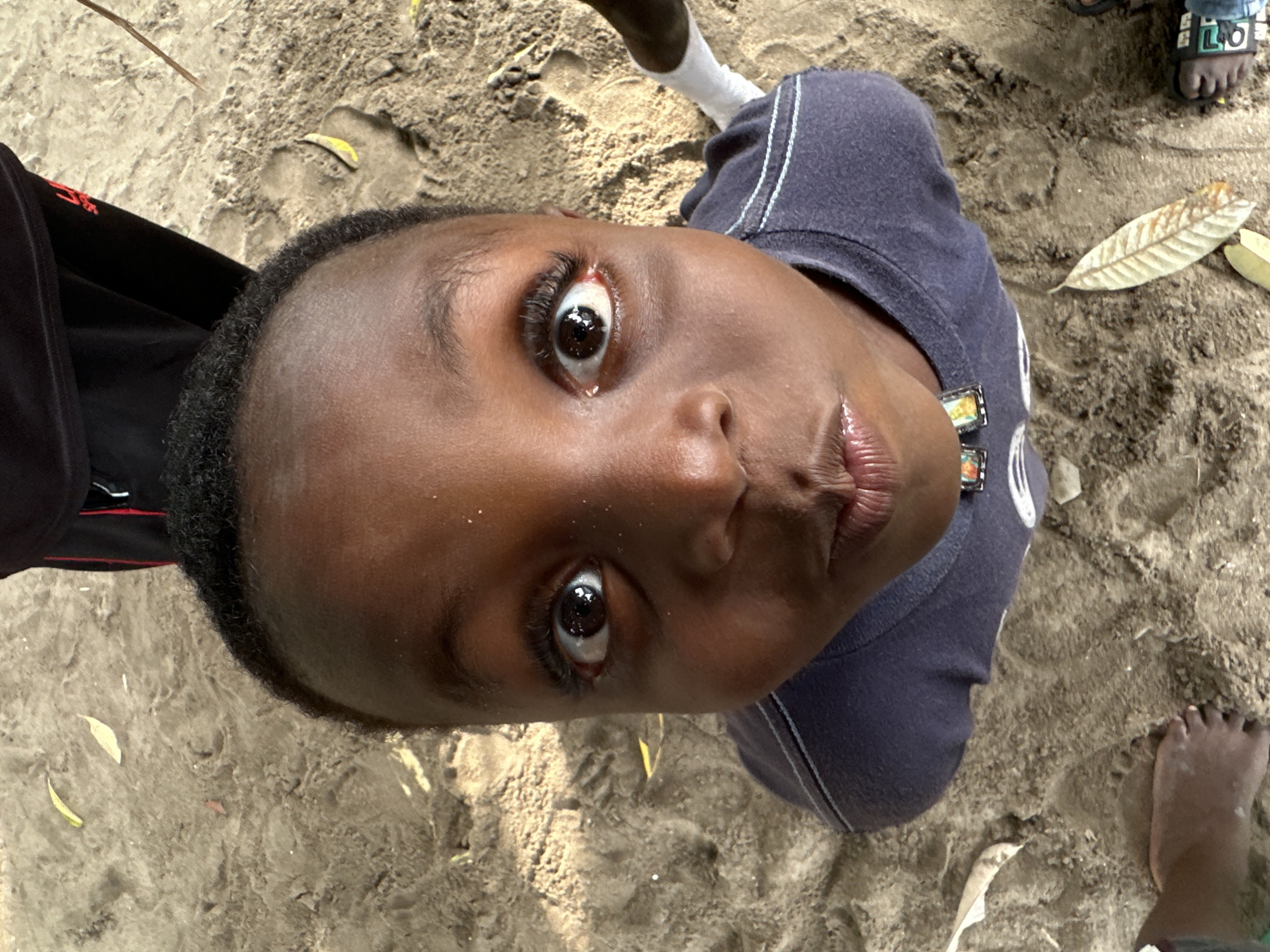 A close-up, high-angle shot captures the face of a young Black child, looking directly into the camera. The child's eyes are wide open, with prominent eyelashes. Their lips are slightly pursed. The child is wearing a dark blue t-shirt with two light blue stripes on the collar and a small emblem on the chest. The background is sandy, with a few fallen leaves scattered across the ground. A portion of black pants with red trim is visible at the top of the frame, suggesting another person is partially in view. The scene appears to be outdoors, possibly on a beach or in a sandy area within Badagry, Nigeria.
