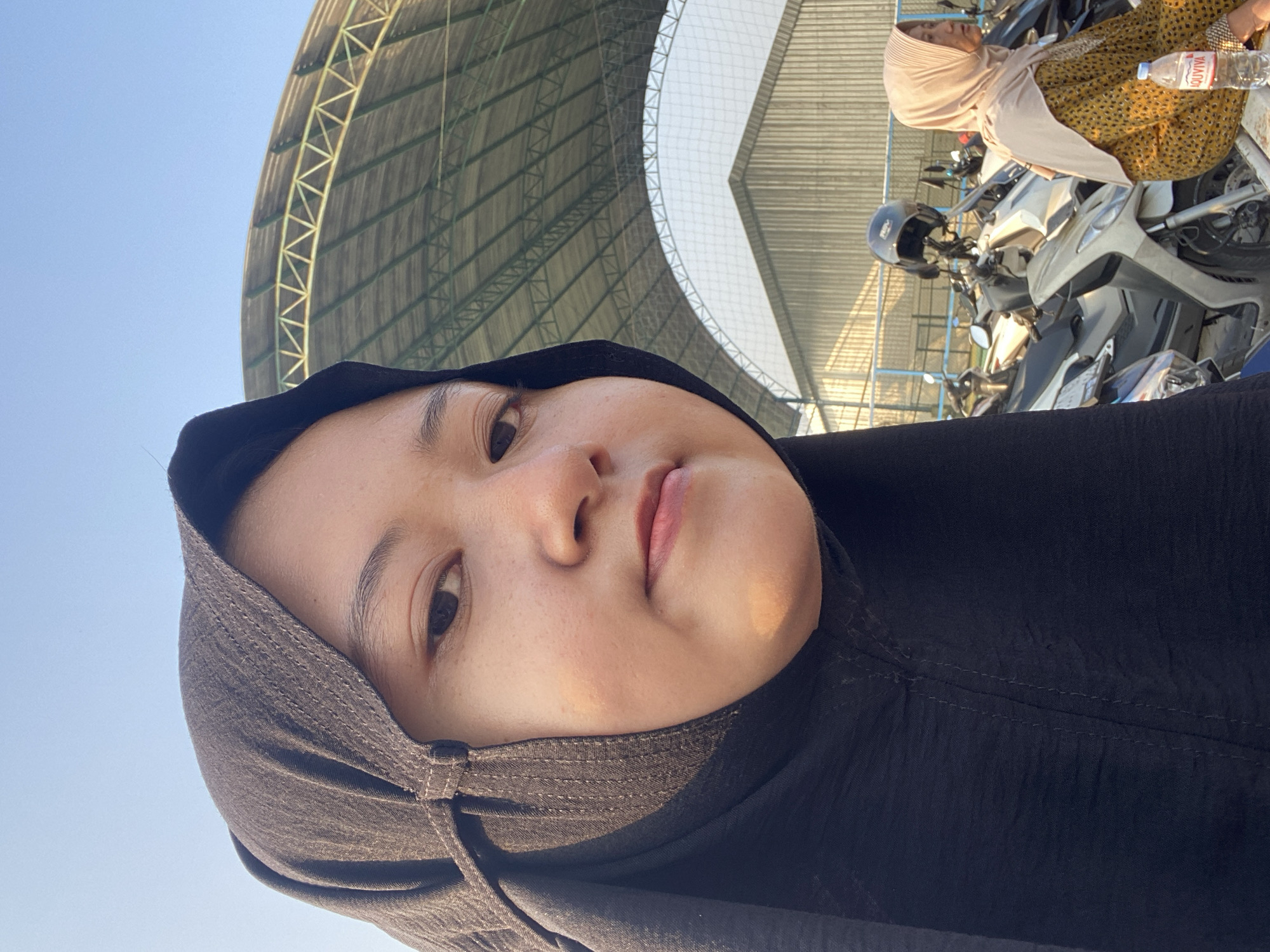 In Sumorame, Indonesia, a woman wearing a black hijab and a patterned yellow dress is visible. The woman is seated and looking to her right, with a bottle of Aquviva water placed on a nearby surface. In the background, a row of motorcycles is parked, with a helmet resting on one of them. Above the motorcycles, a large, arched structure with a metal frame and roof is present, suggesting an outdoor stadium or covered arena. The sky above is clear and blue.