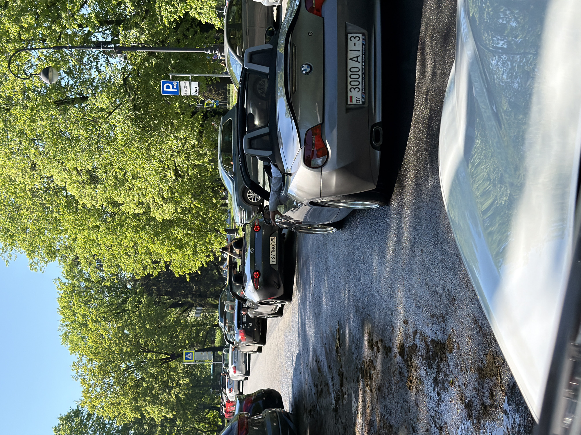 The image depicts a street scene in Saint Petersburg, Russia. A row of parked cars lines the right side of the street, facing the camera. The first car in the row is a gray BMW Z4 with license plate "3000 AI-3" from Belarus. Further down the line, another car has the license plate "Т371НО198."
Street signs are visible on the left side of the street, including a parking sign. Trees with green foliage line the left side of the street as well, obscuring the horizon. The sky is blue, and the asphalt street shows signs of wear.