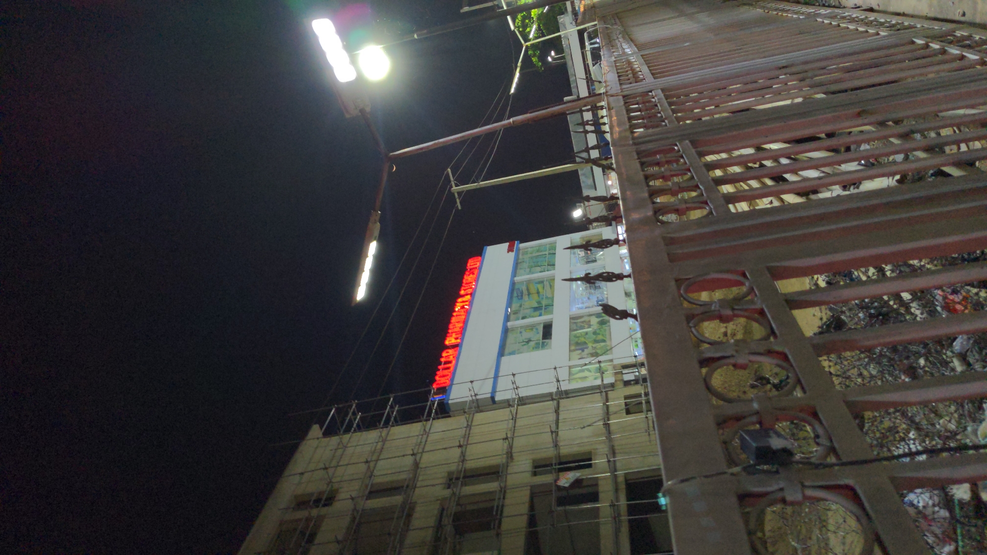 The image shows a building at night in Ado, Nigeria, viewed through a metal gate or fence. The building has a sign in red letters. Scaffolding surrounds the lower section of the building. Above, windows are visible. A street light illuminates the scene from the upper left. The environment is dark, with a dark sky.