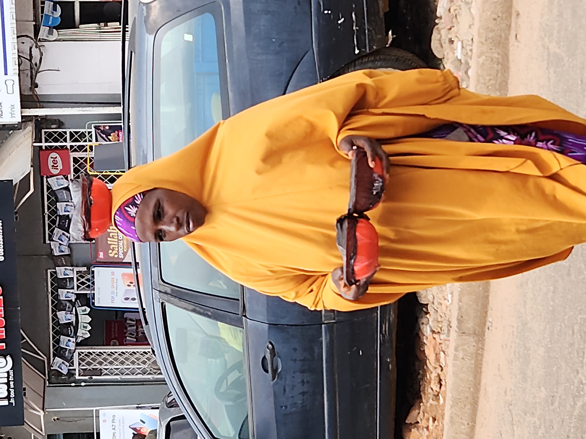 A full-body shot shows a woman in the foreground walking in front of a dark-colored station wagon. She is wearing a bright orange hijab and a matching orange garment that covers her from head to toe. Her face is partially obscured by her hijab. She is holding two red bowls, one stacked on top of the other, in front of her chest. The bowls appear to be empty or contain dark contents. The woman is walking on a dirt and gravel surface, with a sidewalk and a road visible in the background. The station wagon is parked behind her, with its doors closed. A store with an awning and displays of products is visible behind the car. The lighting suggests it is daytime, with some shadows cast on the ground.