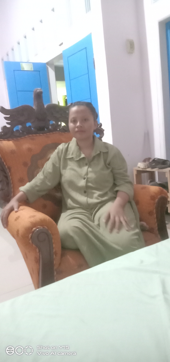 An adult female is depicted seated in an ornate armchair in an indoor setting, identified as Brumbungan Kidul, Indonesia. The female wears a light olive green ribbed collared shirt and matching trousers. Her dark hair is pulled back, and she looks directly at the camera. The armchair features an orange-brown patterned upholstery with a dark, elaborately carved wooden frame, including an eagle sculpture at the top of the backrest. In the background, two bright blue wooden doors are slightly ajar, revealing a darker interior. To the right of the subject, a small light-colored wooden shelving unit holds several dark objects. The room has light-colored walls and appears well-lit. A light-colored surface is visible in the foreground.