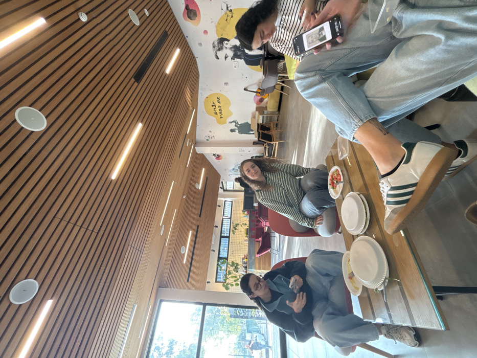 A candid, wide-angle shot captures three young people gathered around a table in a modern, well-lit common area in Kiryat Malakhi, Israel. The image is tilted at a Dutch angle.
The setting features a striking wooden slatted wall and ceiling on the left, integrated with linear strip lights and circular recessed lights. On the right, a white wall is adorned with colorful, stylized murals, including abstract shapes, figures, and a prominent black and white portrait-like depiction of a person. Within a yellow speech bubble on the wall, Hebrew text is visible, reading "אנשים הזזים את" (Anashim Ha-Zazim et), which translates to "People who move the..."
Three individuals are seated at a wooden table with a glass top. In the foreground, a person wearing glasses and a dark jacket holds an object in their hands. Across from them, another person with curly hair, wearing a striped long-sleeve shirt and light pants, smiles faintly towards the camera. On the far side of the table, a third person, partially visible, wears a striped shirt and light blue jeans, with white and green Adidas sneakers. They are looking at a smartphone, which displays a black and white image of a person.
Several white paper plates are on the table, some containing food, possibly salad. Large windows along the back wall let in bright natural light, indicating it is daytime, and offer a glimpse of green trees outside. The space appears to be a casual lounge, cafeteria, or community center, designed for social interaction.