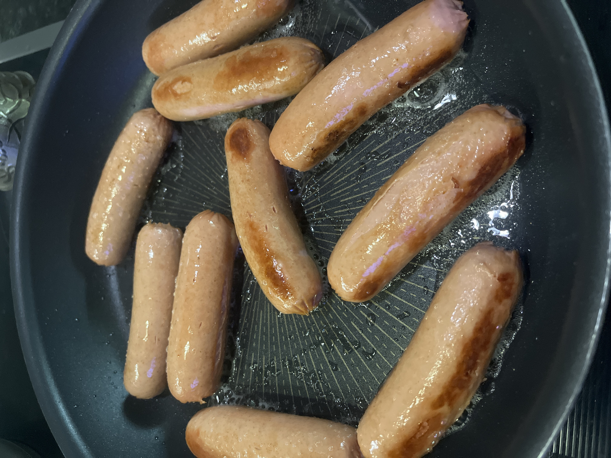 A top-down shot shows several cooked sausages arranged in a black frying pan. The sausages vary in color from a pale pink to a browned tan, indicating varying degrees of cooking. Traces of oil or grease are visible in the pan, suggesting they were fried. The setting appears to be a kitchen. The image provides no evidence to pinpoint the location as Krasnodar, Russia.