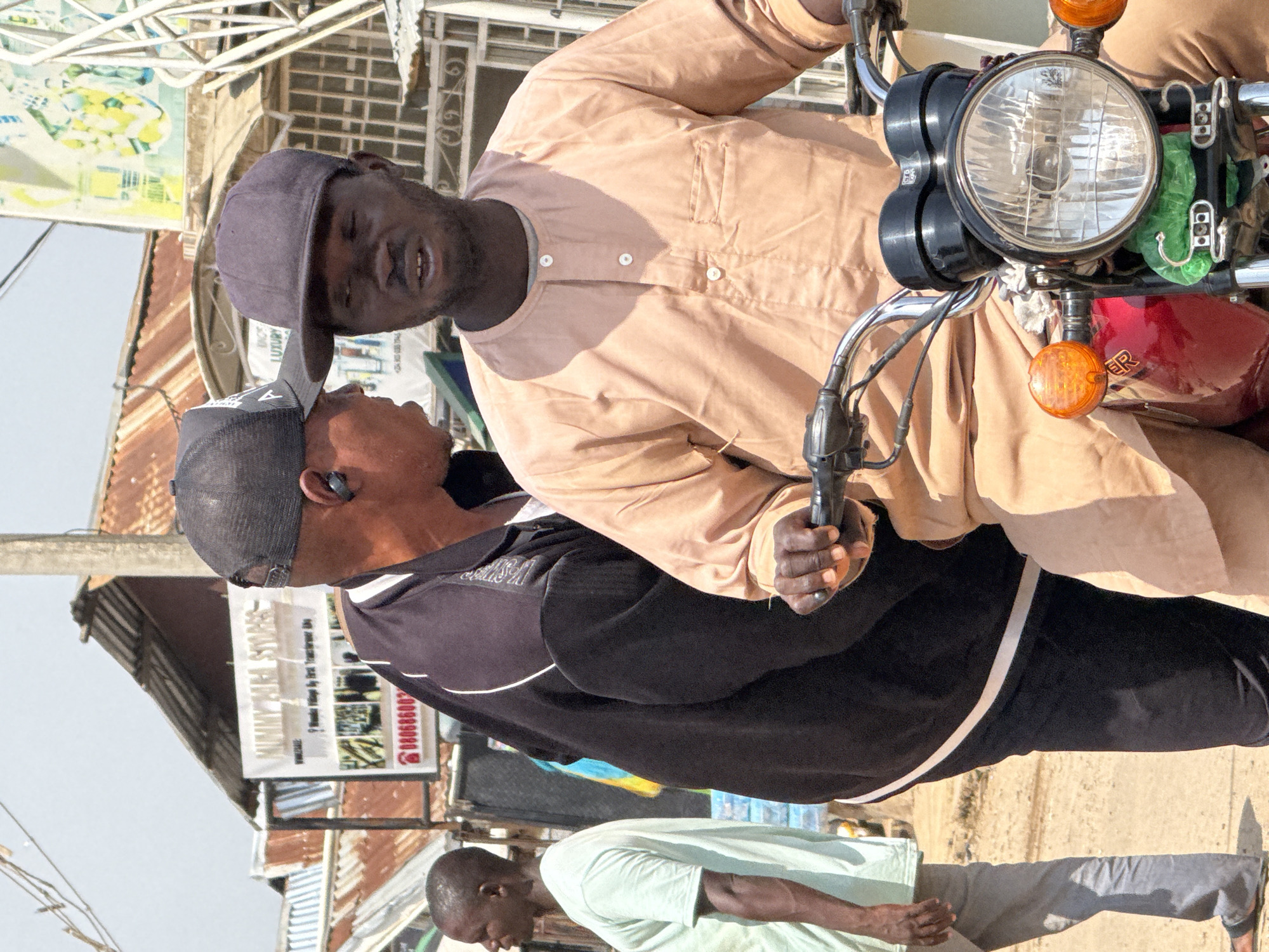 This image captures a street scene in Abuja, Nigeria. In the foreground, two men are interacting. The man on the right is seated on a motorcycle, wearing a light brown, long-sleeved garment with white buttons and a cap. He is looking forward with his mouth slightly open. The man on the left is standing and facing the seated man, wearing a black jacket with white trim and a black baseball cap. He is also looking at the seated man and appears to be speaking, with an earbud visible in his left ear. A third man, partially visible on the left side of the image, stands facing away from the camera, wearing a light green t-shirt and grey pants. The background shows a bustling urban environment with buildings, corrugated metal roofs, and signs, suggesting a market or commercial area. One sign prominently displays "MUMMY MIYA STORES." The ground is dusty and appears to be unpaved. The lighting suggests it is daytime, likely under a bright sun, creating shadows.