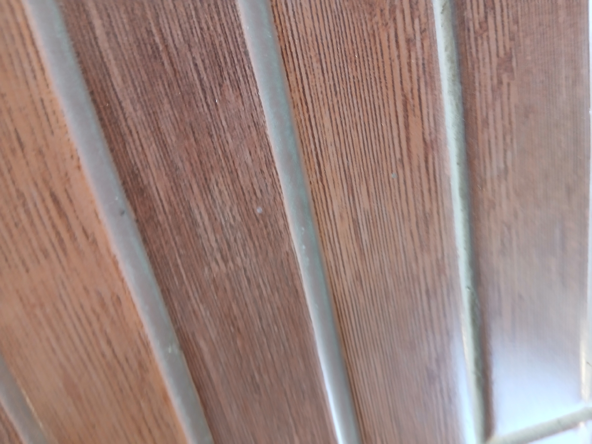 The image shows a close-up, oblique view of a wooden surface with vertical grooves. The wood appears to be a warm brown color with a visible grain pattern. There are distinct, slightly raised or indented lines running vertically across the surface, creating a ribbed or paneled effect. The lighting suggests a natural light source, casting subtle shadows along the grooves. No people, animals, or specific identifiable landmarks of Sekarputih, Indonesia are visible. The scene is purely focused on the texture and pattern of the wooden surface.