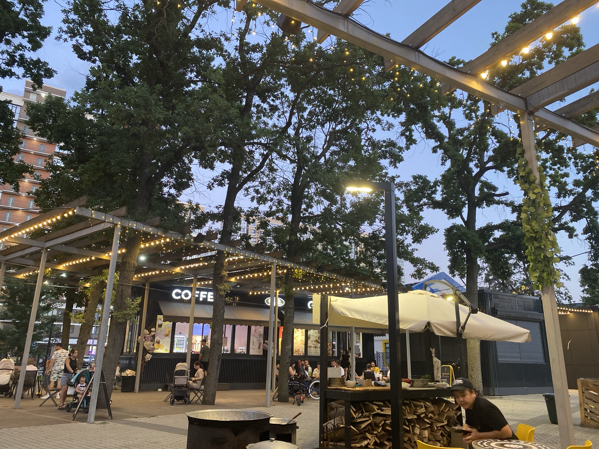 This image depicts an outdoor seating area of a cafe called "COFFEE" in Krasnodar, Russia, during the evening. The area is covered by a wooden pergola adorned with string lights. Several trees are present, with their branches extending through the pergola. In the background, a multi-story brick building is visible.
There are multiple people in the scene, some seated at tables and others standing. A woman and a young child are in the foreground, with a stroller nearby. Another woman is seated at a table with a stroller. A man is seated at a table in the right foreground, looking down at his phone. Other patrons are visible in the background, some eating and drinking.
Objects in the scene include tables, chairs, strollers, a trash can, a stack of firewood, and outdoor cooking equipment. The lighting suggests it is dusk, with the string lights and a street lamp illuminating the area. The ground is paved with interlocking bricks. The overall atmosphere appears casual and relaxed.