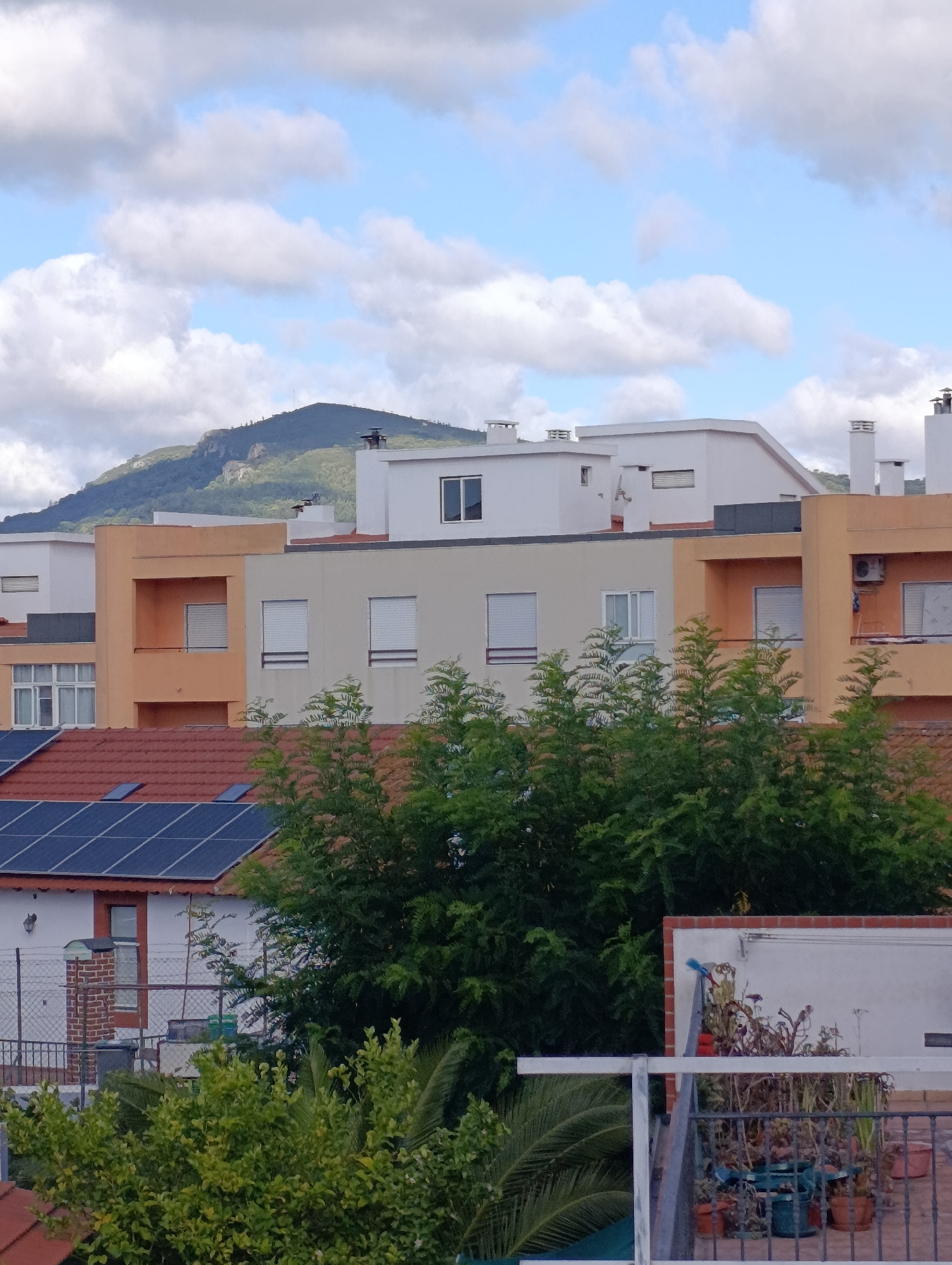 The image shows a view of buildings and a hillside under a partly cloudy sky. The buildings are of various heights and colors, with flat roofs, some featuring chimneys or air conditioning units. Solar panels are visible on the roof of a lower building in the foreground. A lush green hillside with trees and rocky outcrops forms the background, with a distant mountain peak visible at the top. The scene appears to be a residential area. The lighting suggests it is daytime.