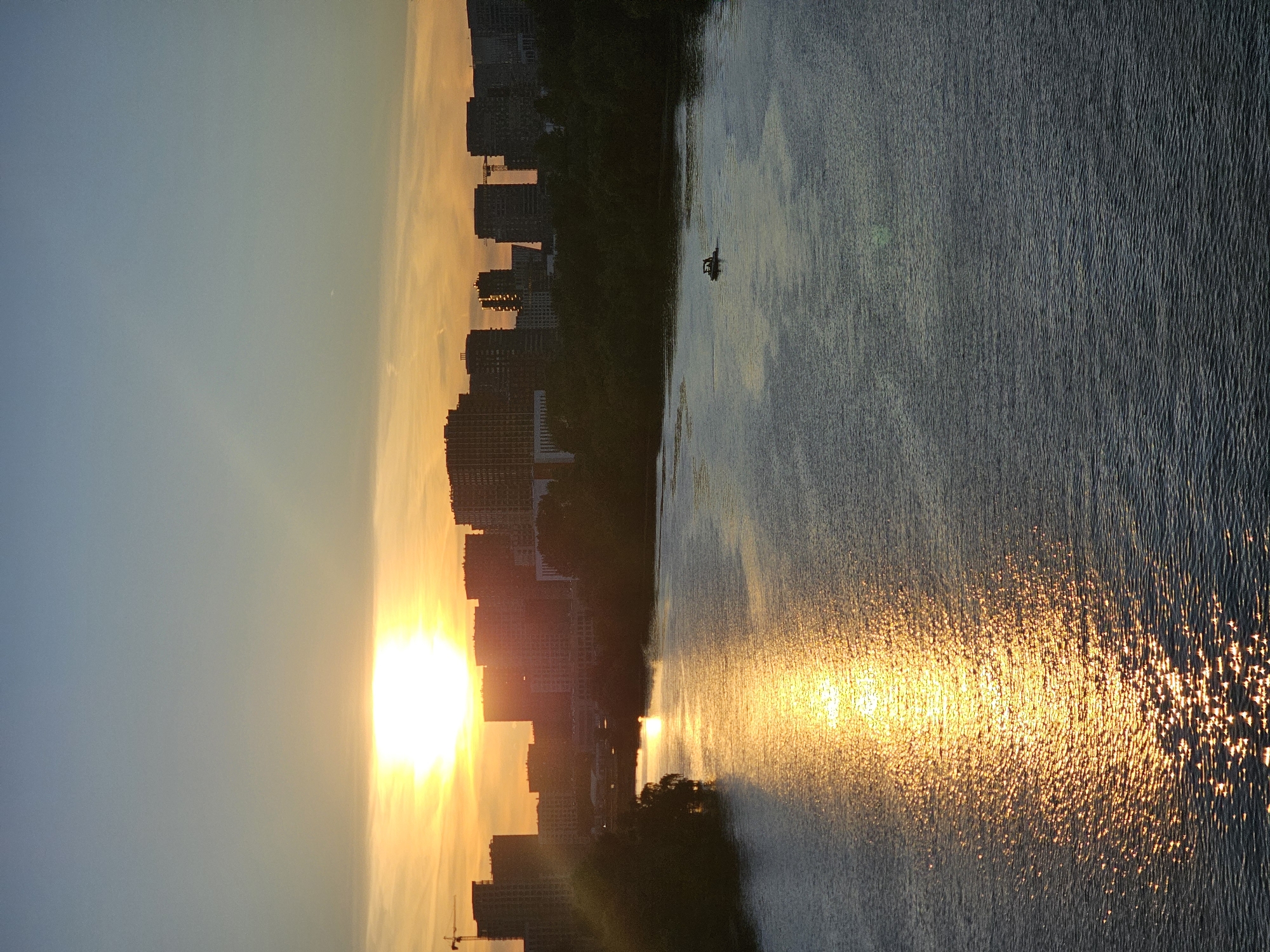 This is a photograph of a sunset over a body of water, likely a river or lake, with a cityscape in the background. The sun is low on the horizon, casting a warm, golden light across the scene. The water reflects the vibrant colors of the sunset, creating a shimmering pathway of light.
In the foreground, the water is textured with small ripples, catching the sunlight and creating a sparkling effect. A small boat with two figures is visible on the water in the middle distance. Behind the water, a dark, silhouetted line of trees and vegetation separates the water from the urban landscape.
The cityscape consists of numerous tall buildings, appearing as dark, angular shapes against the bright sky. Some of the buildings seem to be under construction, with cranes visible. The sky transitions from a soft blue at the top to a warm orange and yellow near the horizon, indicating the time of day is sunset. The overall mood is tranquil and picturesque, with the warm light evoking a sense of peace and beauty.
The location is identified as Khimki, Russia. No visible text is present in the image.