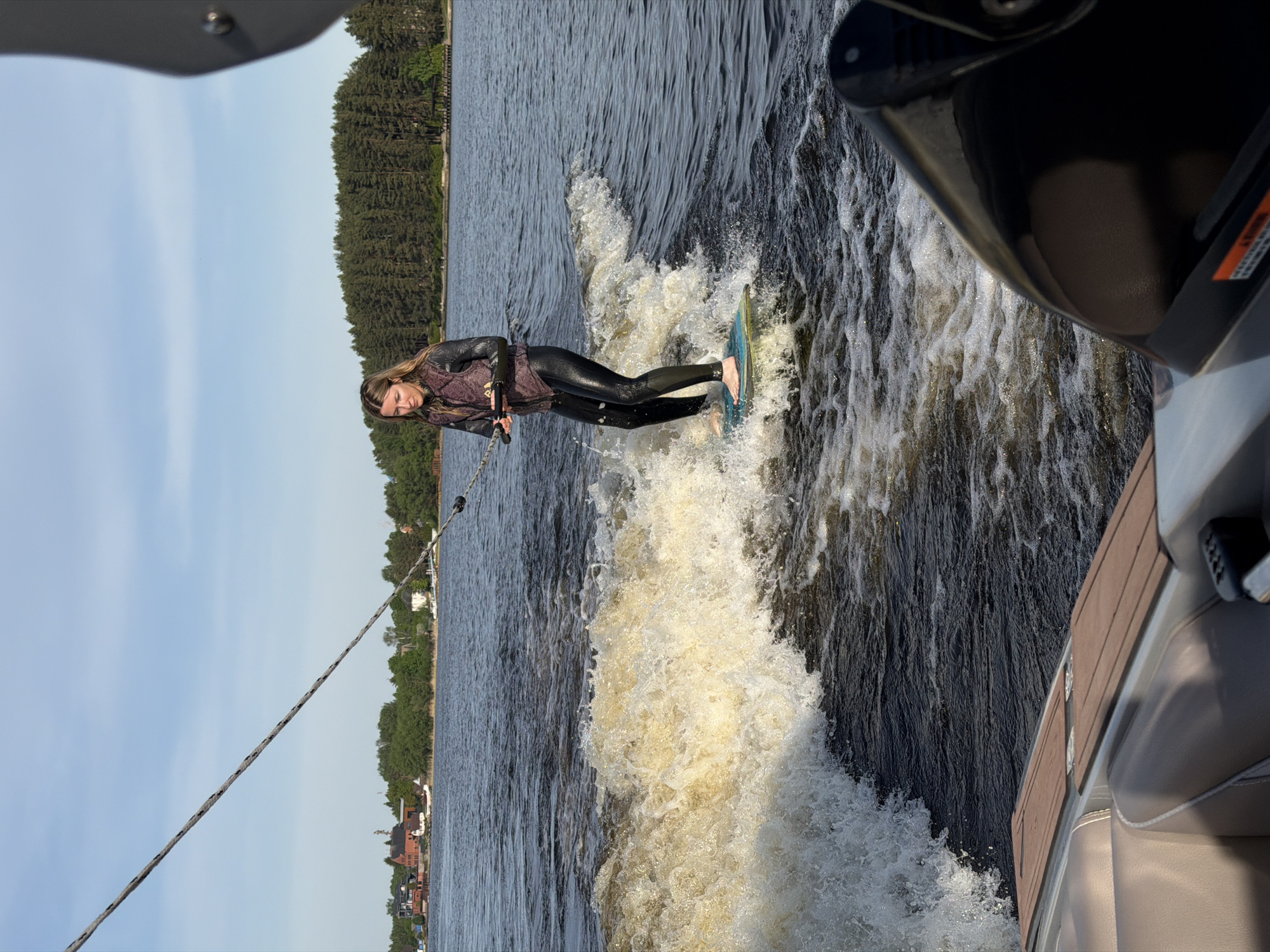 A woman is wakeboarding on a body of water in Терехово, Russia. She is wearing a wetsuit and is being pulled by a rope attached to a boat. The boat is visible in the right of the frame, and trees line the shore in the background. The sky is a pale blue. The woman is standing on the wakeboard with her feet bare.