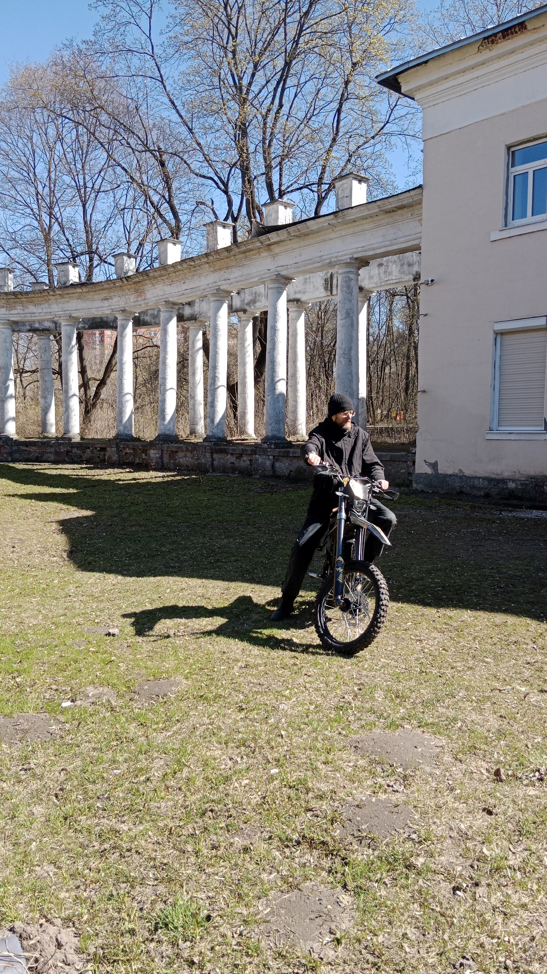 The photograph captures a man seated on a motorbike in an outdoor setting. The location is identifiable as Balashikha, Russia, specifically in front of the estate building at st. Zarechnaya, 16. The man is wearing a dark beanie, glasses, and a black jacket. He is positioned on a motorbike, facing the camera. Behind him, there is a classical colonnade structure, along with a building with closed shuttered windows. The ground is covered in grass, with visible shadows cast by the man, motorbike, and colonnade. The trees in the background are bare, suggesting it is either early spring or late autumn. The sky is clear and blue. The overall scene suggests a casual moment with historical architecture in the background.
