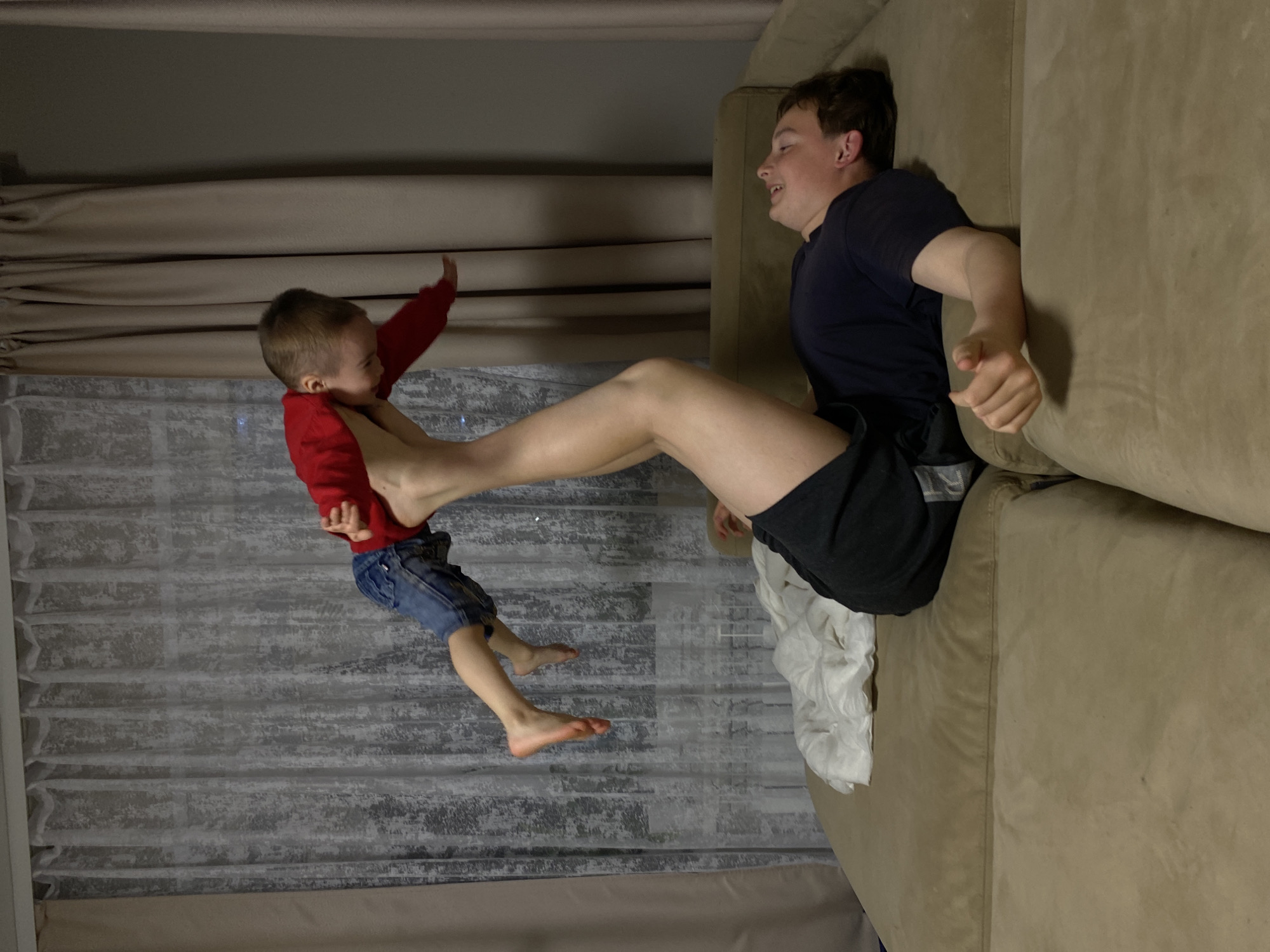 A photograph depicts a young boy in a red shirt and blue shorts being held in the air by the foot of a teenage boy lying on a tan couch. The younger boy's facial expression is unclear. The teenage boy is wearing a dark blue shirt and black shorts, and he appears to be laughing. Behind them is a window covered with gray and white curtains. The location is suggested to be in the city of Ольховка, Russia.
