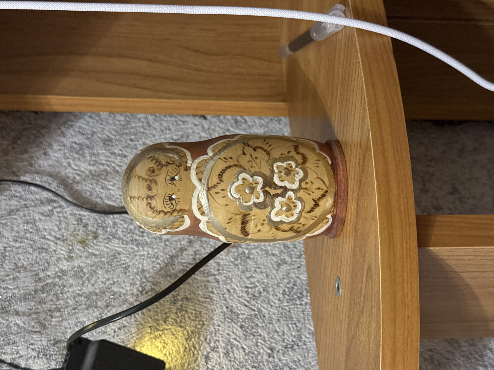 A single Matryoshka doll, painted with an intricate floral design and a stylized face, rests on a wooden shelf. The doll is brown and tan, with white accents on the flowers and face. To the left of the doll, a black electrical cord is visible, attached to a black power adapter that emits a soft yellow light. The background is a textured, gray wall, possibly a carpet or fabric. To the right of the doll, a white cord runs vertically, and a pen with a clear plastic tip is lying on the shelf. The shelf itself is made of polished light brown wood, with a visible metal screw in the center. The overall scene appears to be indoors, likely a domestic setting. This scene is located in Масаловка, Russia.