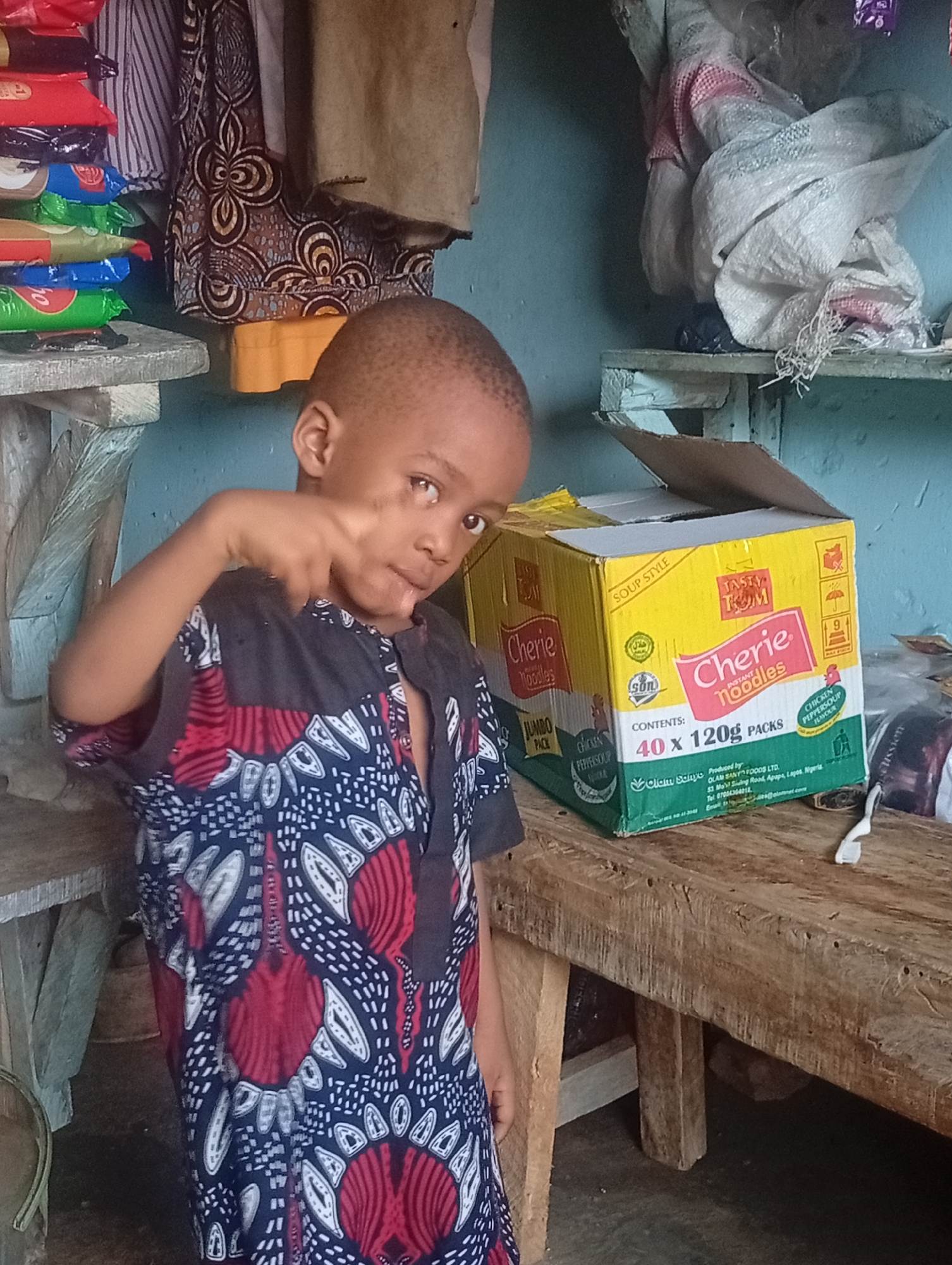 A young boy wearing a patterned blue, black, and red shirt is standing in what appears to be a small shop or kitchen. The boy is making a gesture with his hand, holding up his index and middle fingers. To his right is a box of "Cherie Noodles" instant noodles, partially opened. The box is yellow, green, and white.
Behind the boy, there is a cluttered environment. A shelf to the left holds stacks of packaged goods, while another shelf above and to the right has a white bag or cloth. The walls are painted a light blue color. A wooden table is to the right of the boy and the noodle box.