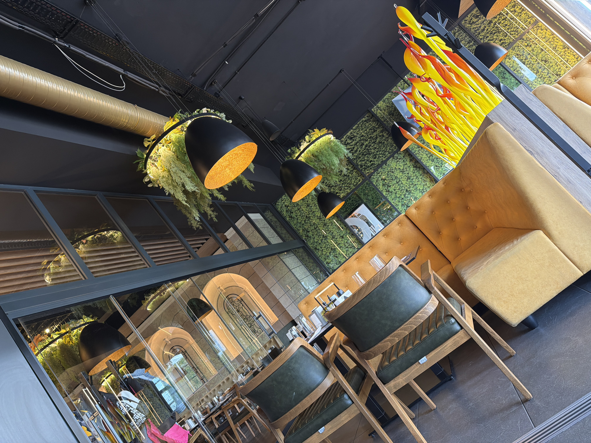 The image depicts the interior of a restaurant in Burgas, Bulgaria. The design features a modern aesthetic with elements of greenery. The interior includes tables and chairs, a cushioned seating area, and light fixtures. The decor incorporates decorative plants, and there is an art installation featuring yellow and red elements. The setting appears to be a commercial space with a focus on creating an inviting and contemporary atmosphere.