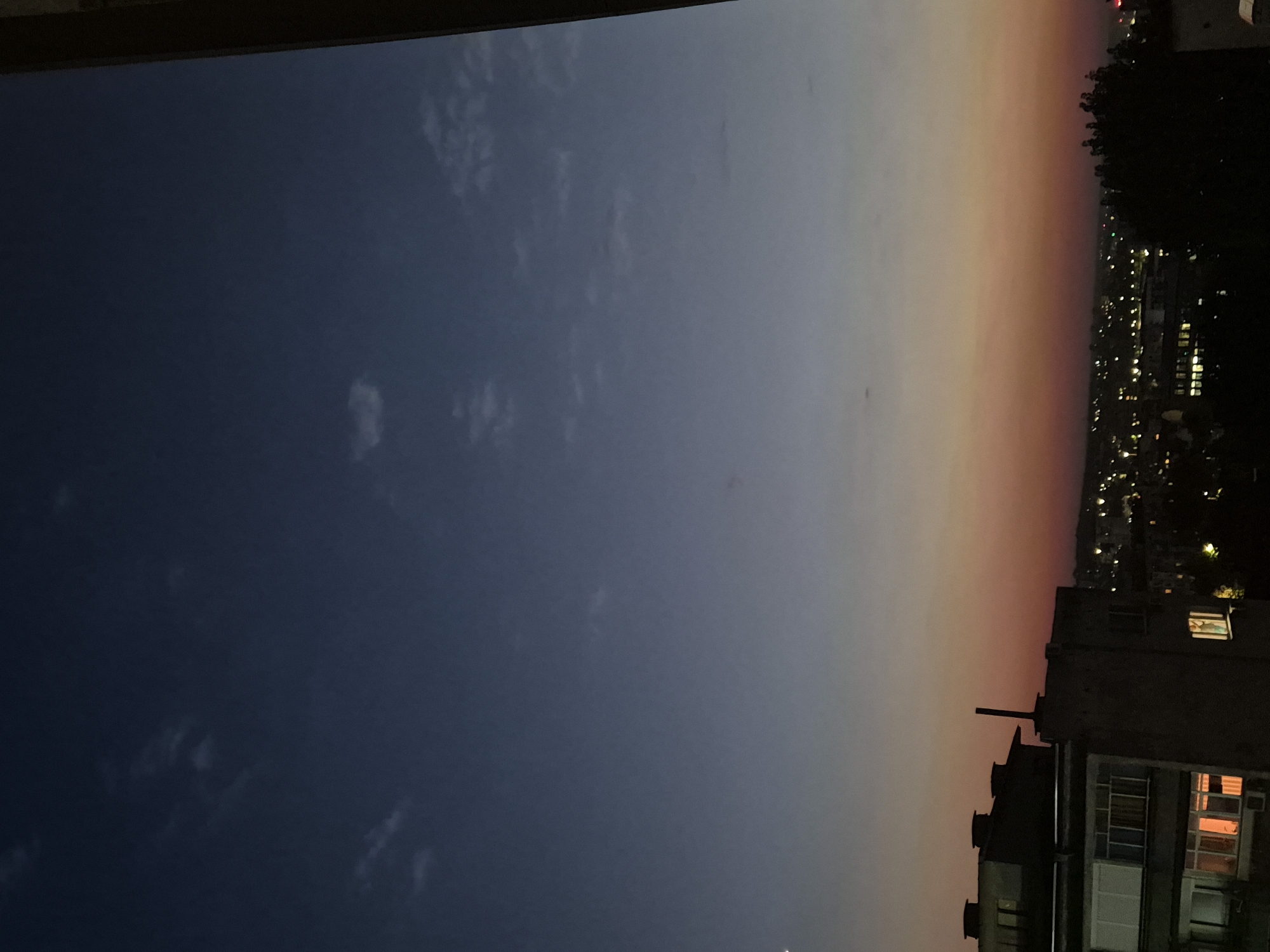 The image shows a cityscape at dusk, with a gradient of colors in the sky transitioning from deep blue at the top to orange and pink near the horizon. Silhouetted against the sky are buildings, some with lights on, and trees. The foreground features a closer view of a building with several windows, some of which are illuminated. The overall impression is of a quiet evening in a city.